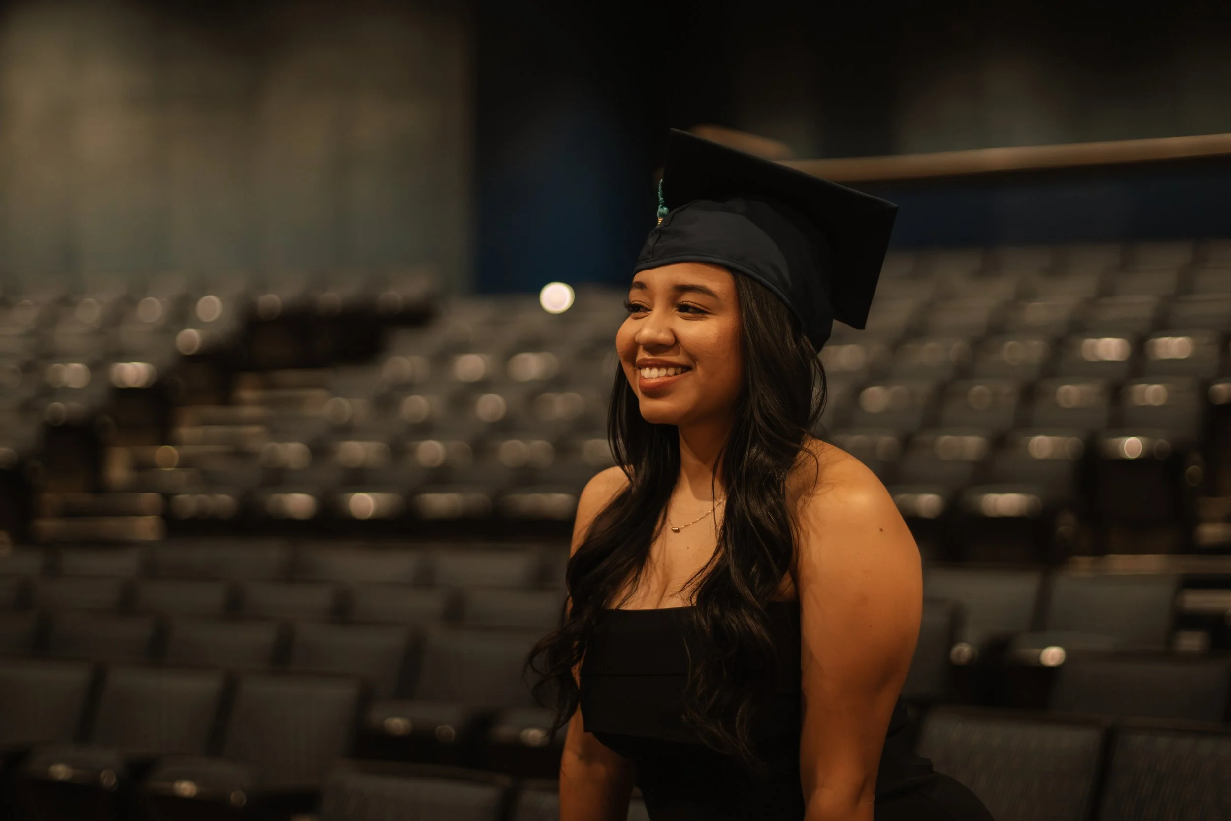 A young woman in a graduation cap and gown smiles while standing in an empty auditorium or stadium.