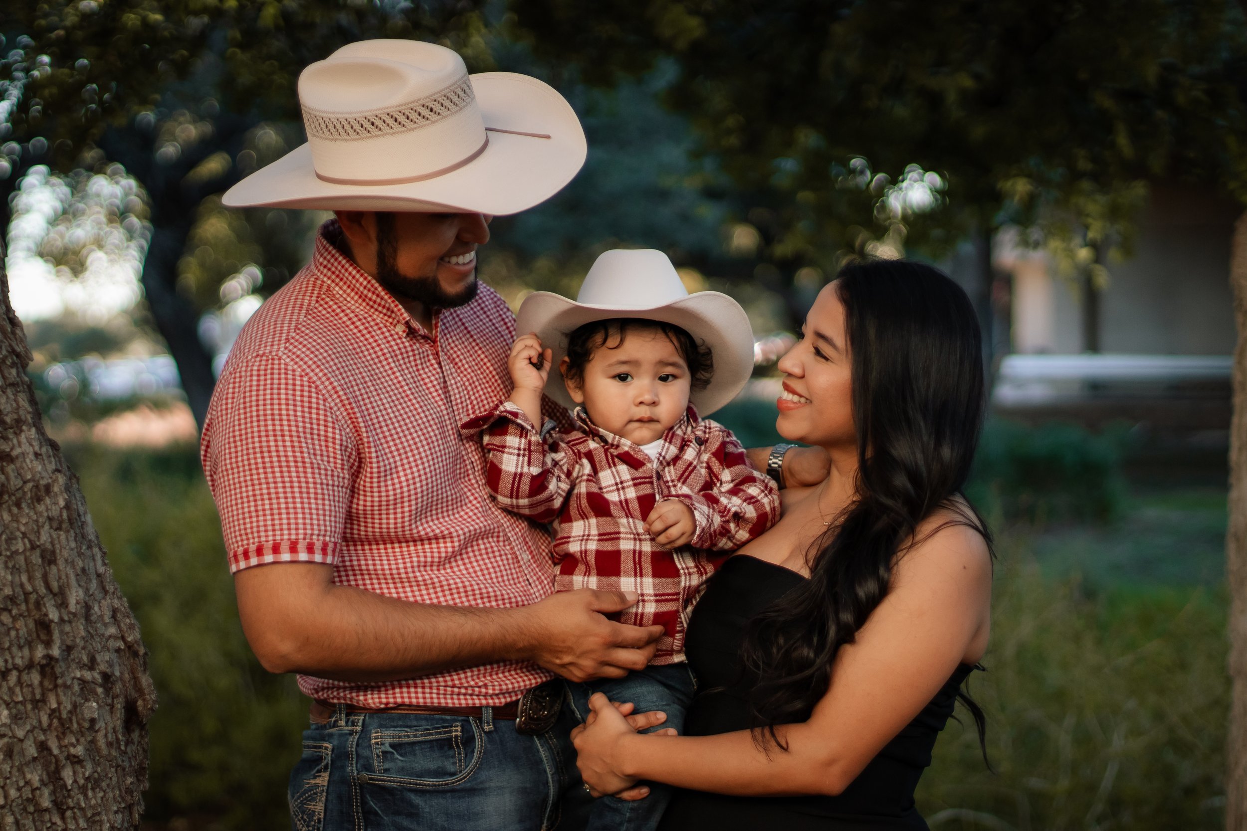 A family of three enjoying an outdoor moment, with the father and son wearing cowboy hats and matching red plaid shirts, and the mother in a black strapless dress, all smiling and looking at each other, surrounded by trees and natural scenery.