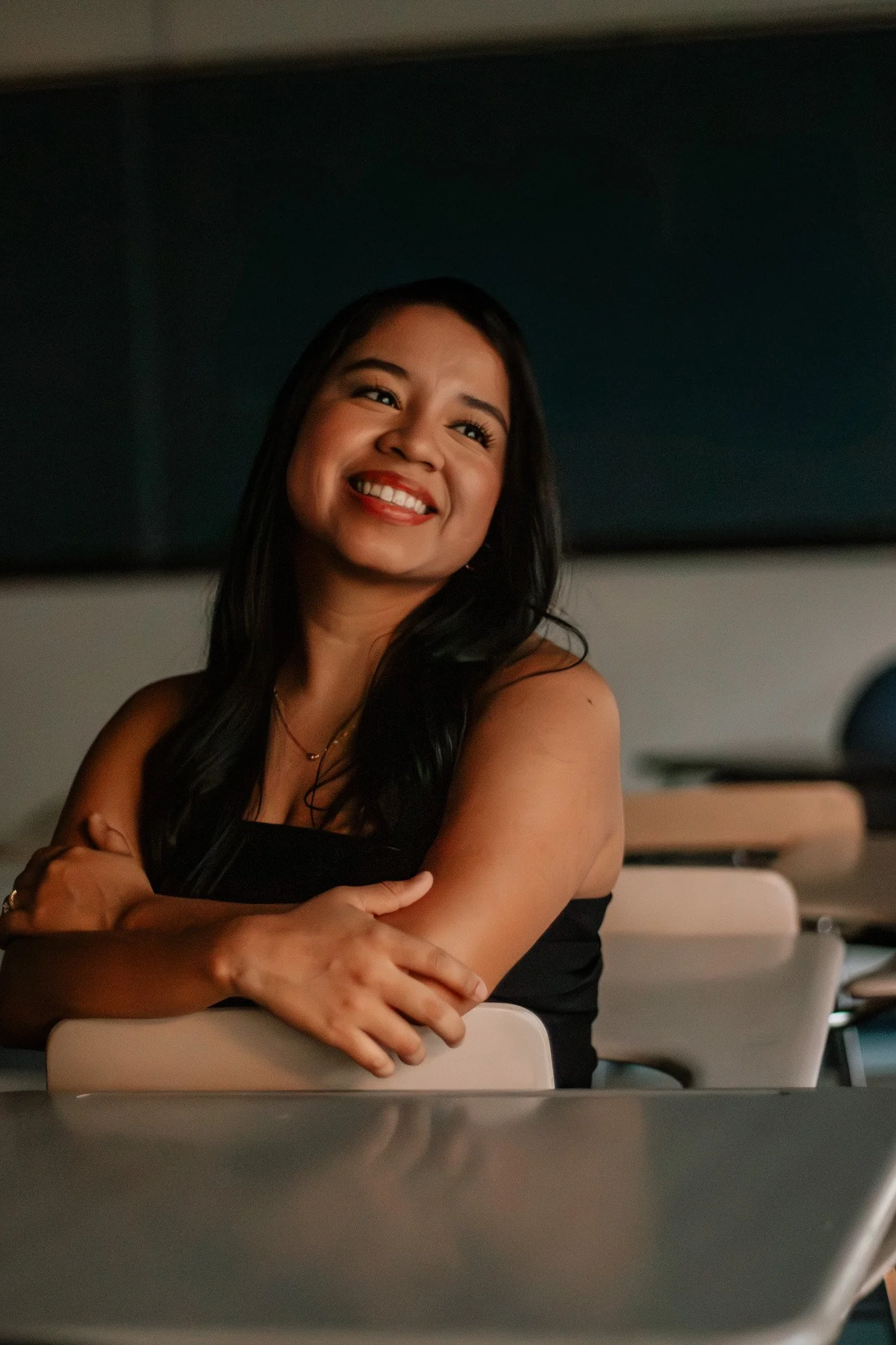 A woman with long black hair smiling and looking to her left, sitting at a desk or table with her arms crossed.