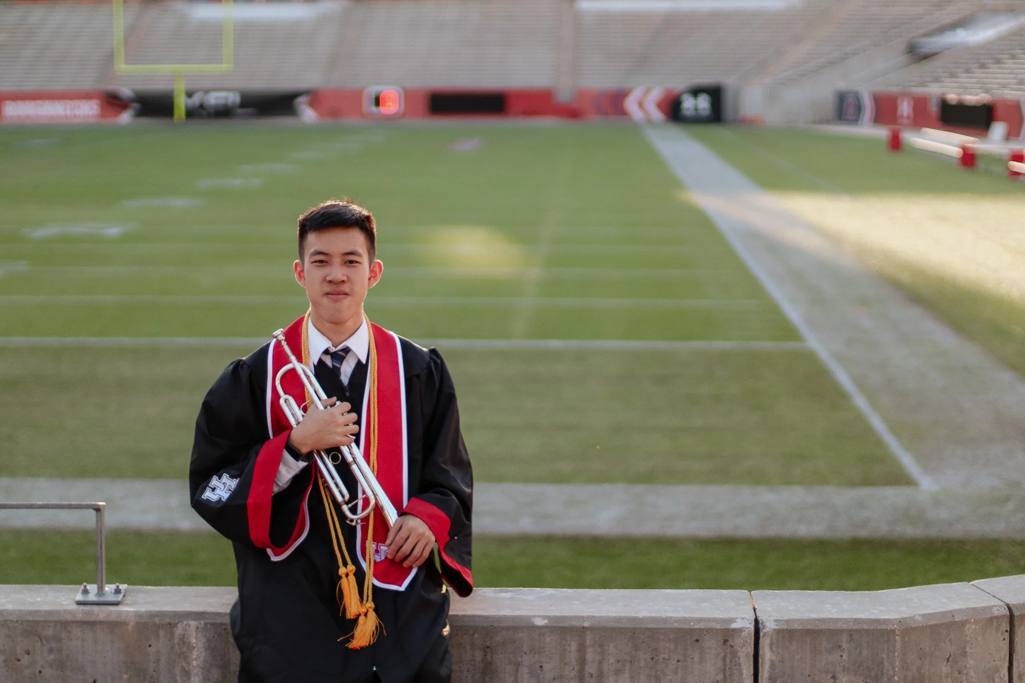 A young man in graduation gown and tie holding a trumpet, standing on a stadium step with an empty football field in the background.