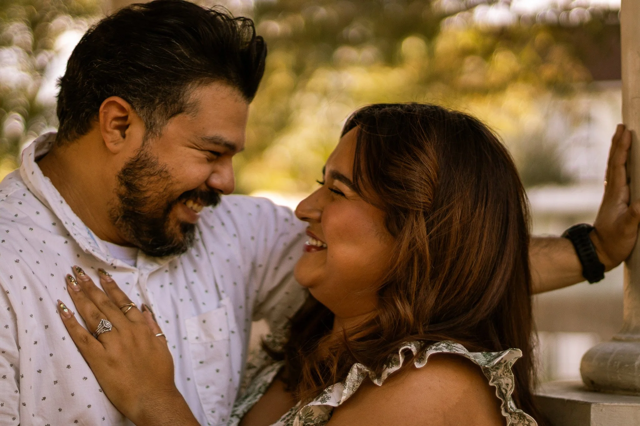 A man and woman are close, smiling and touching foreheads, outdoors with blurred trees in the background.