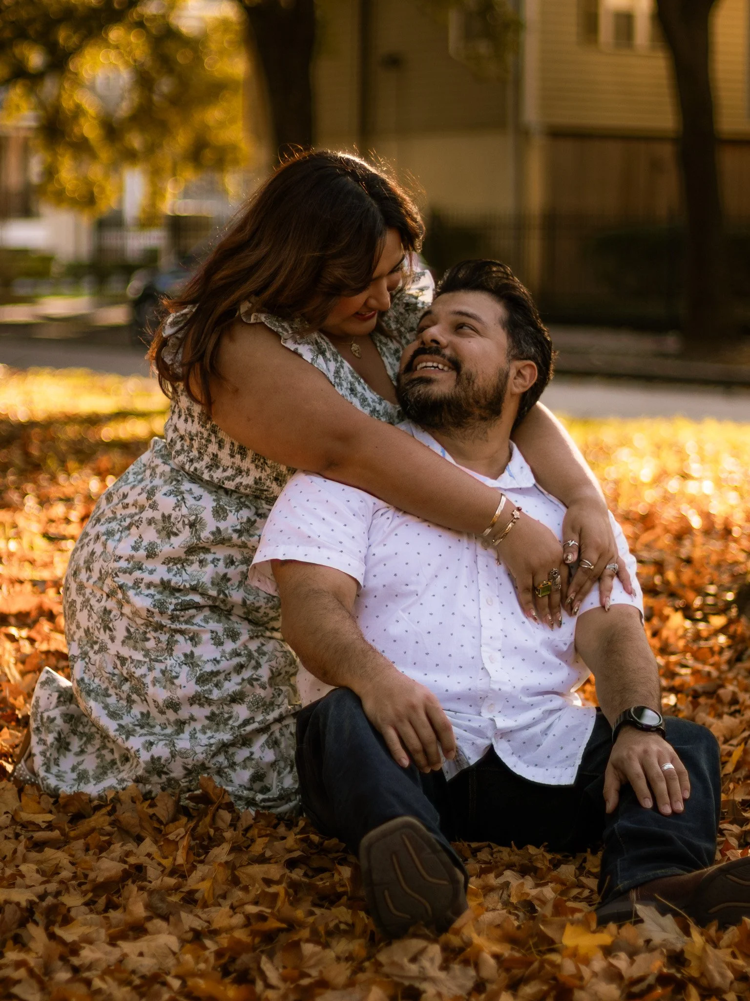 A loving couple sitting on autumn leaves in a park, smiling and sharing an embrace during golden hour.