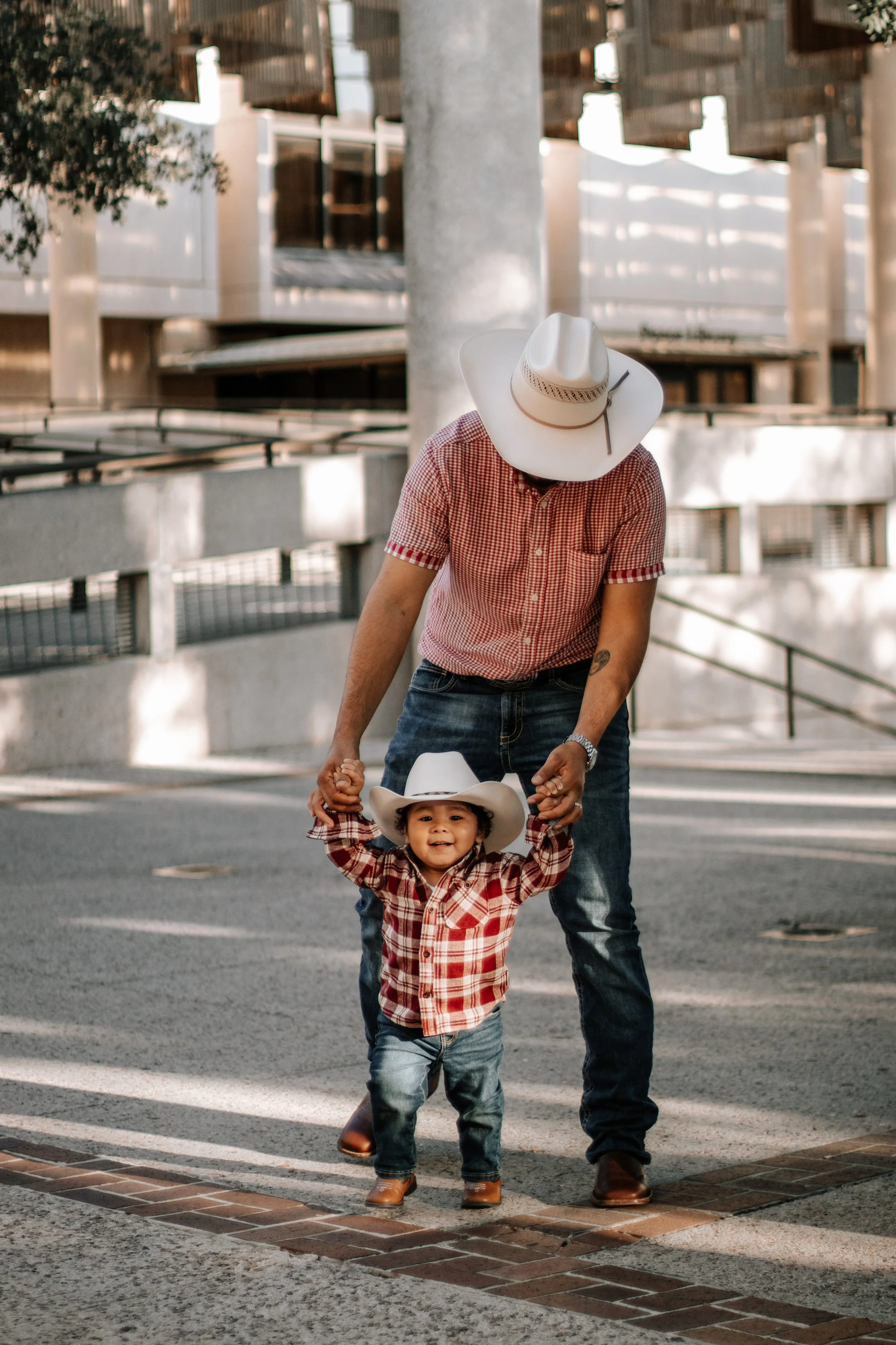 A man and a young boy in cowboy hats and western attire, outside on a city street, holding hands, with modern buildings in the background.