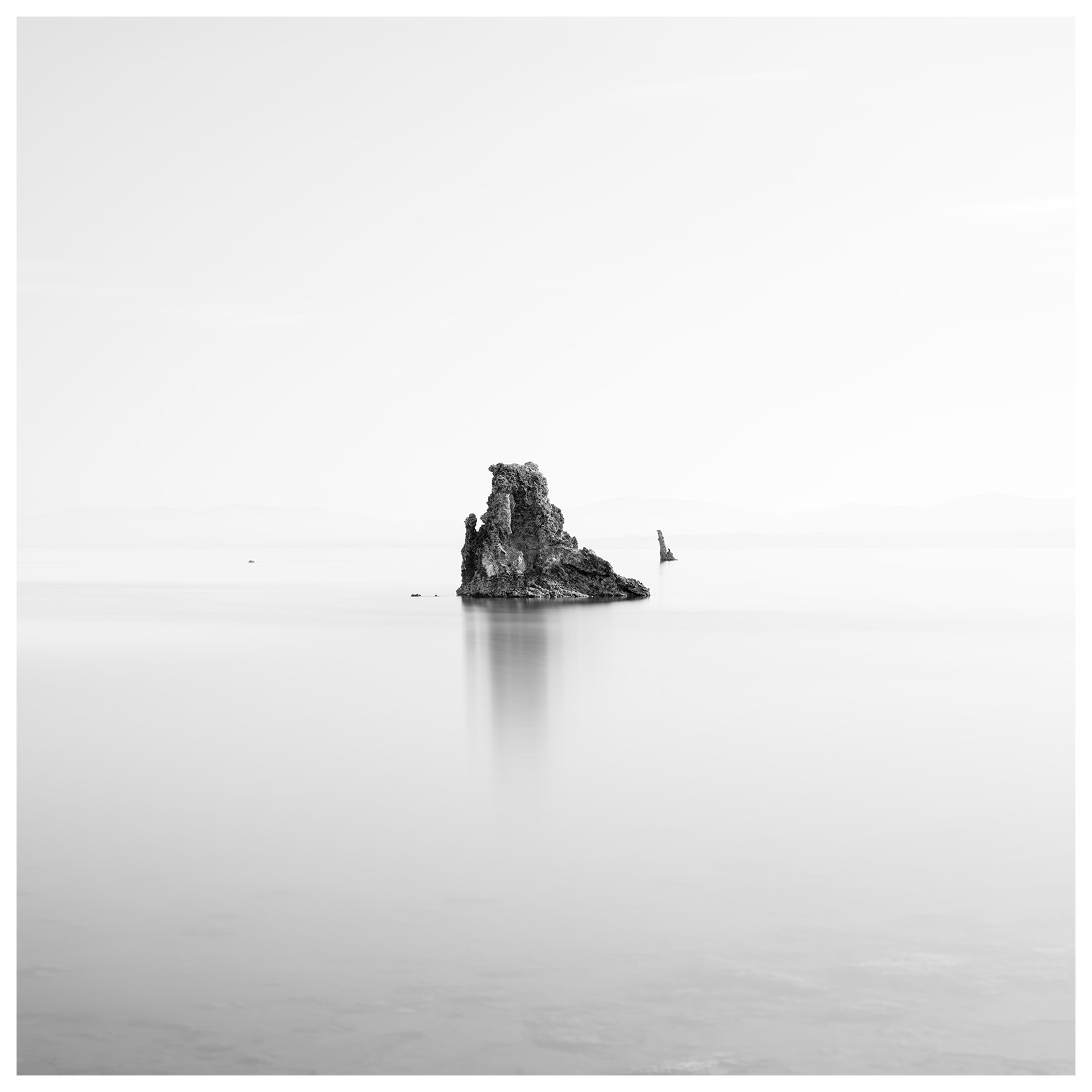 A black and white photograph of a large rock formation in calm water with a lighthouse in the distance.