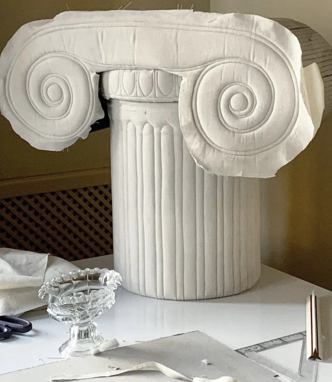 A large, white marble sculpture of an Ionic column capital, including volutes. The sculpture appears to be in the process of being carved or assembled, with tools and materials surrounding it.