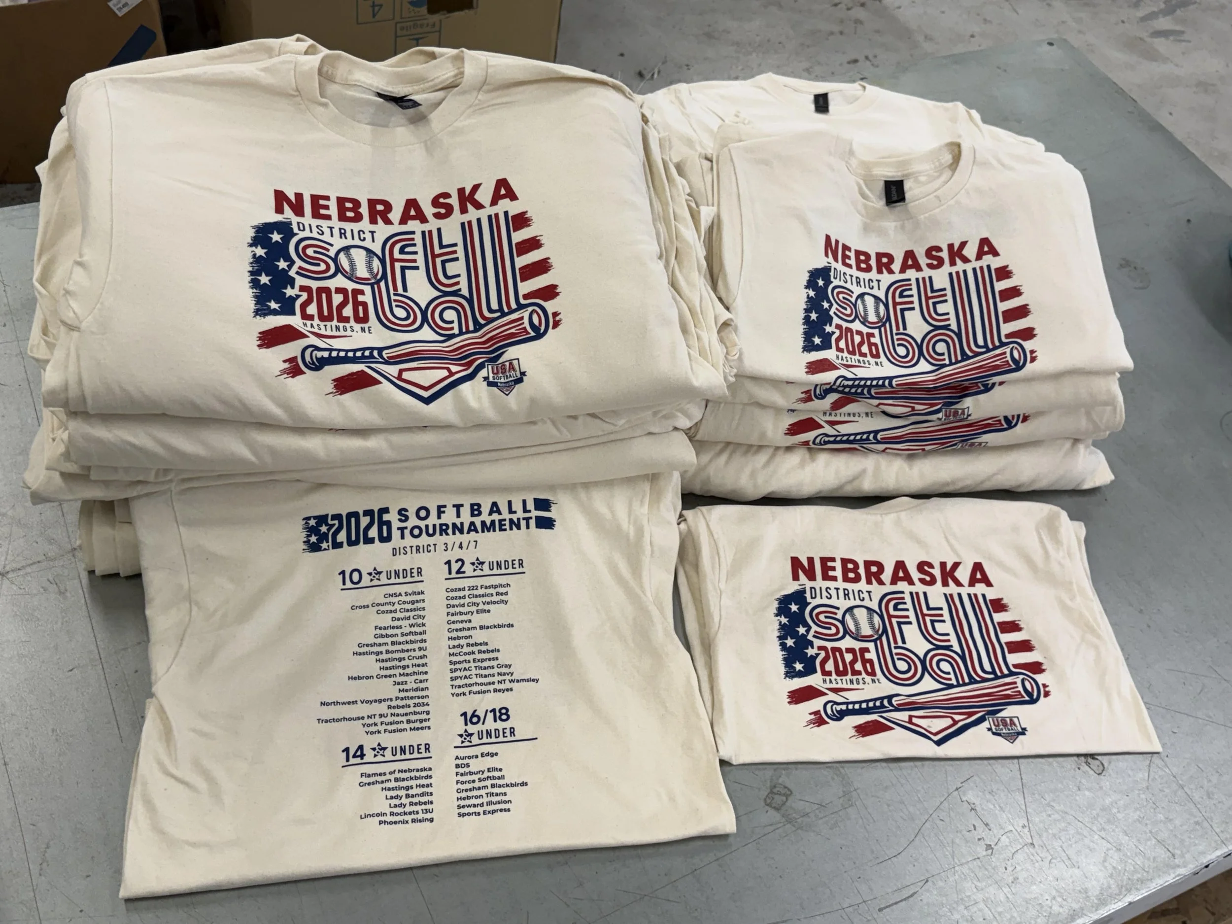 Stacked white T-shirts with red, blue, and black printing advertising the Nebraska Softball District Tournament 2026, featuring a baseball bat, American flag, and list of participating teams.