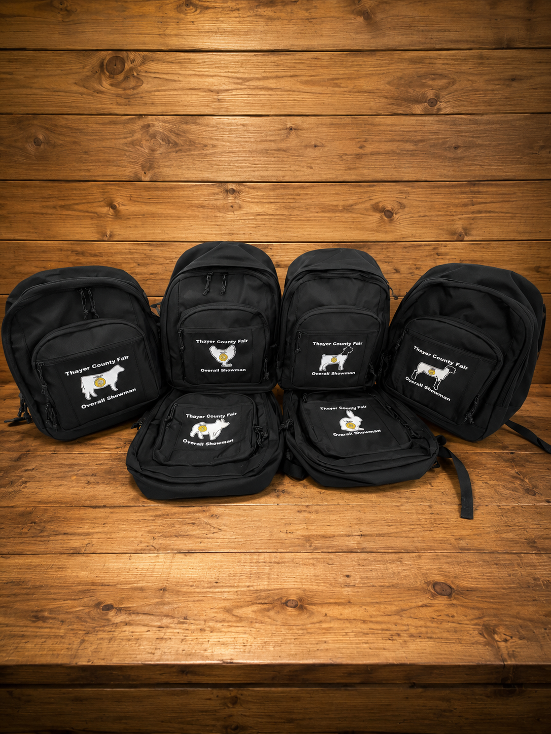 Black DTF backpacks with Thayer County Fair logo, featuring Different animals, displayed on a wooden surface against a wooden wall.