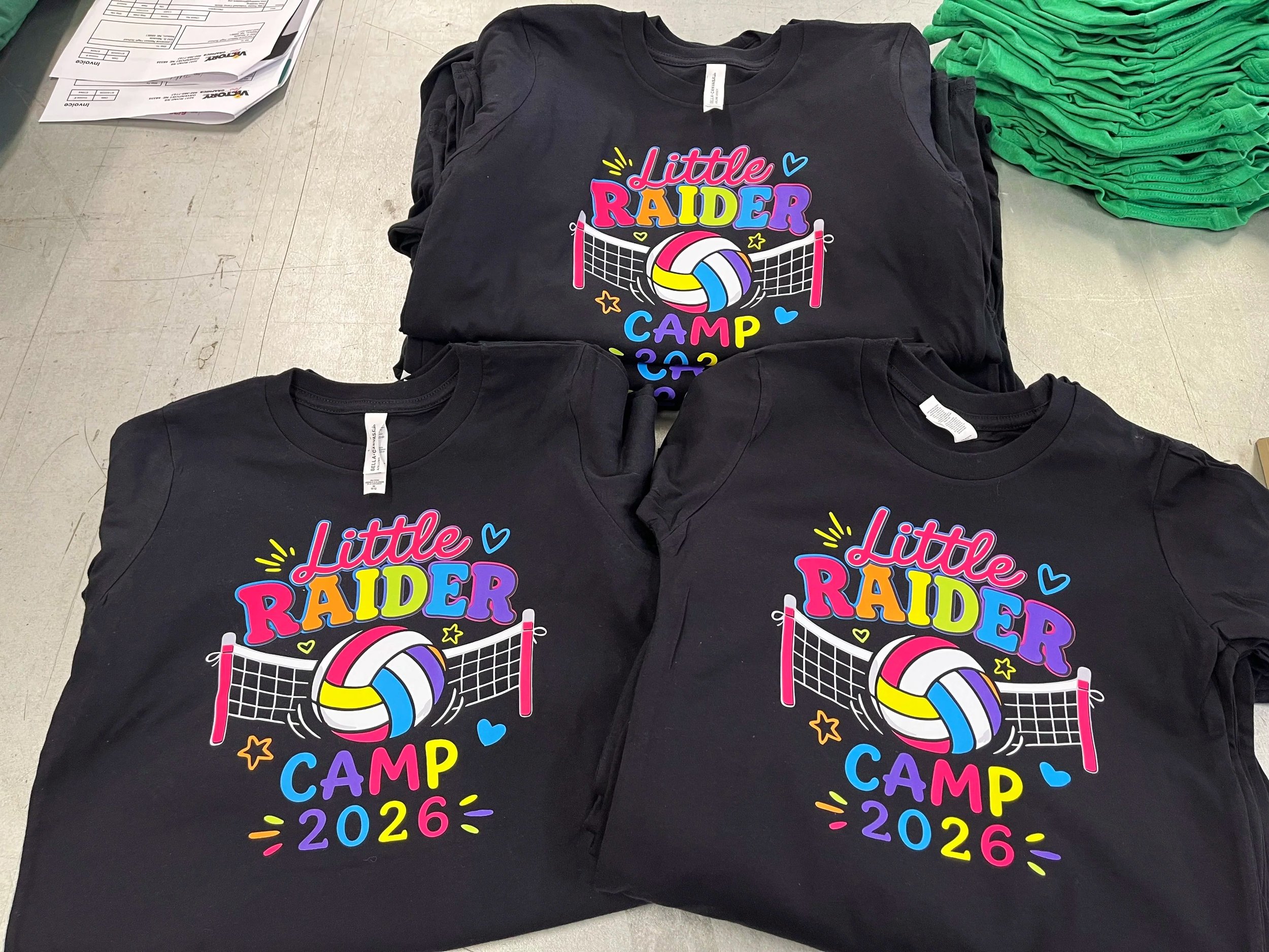 Stacked black DTF T-shirts with colorful 'Little Raider Camp 2026' graphics, featuring a volleyball, a net, and stars on a table alongside green folded shirts and some papers.