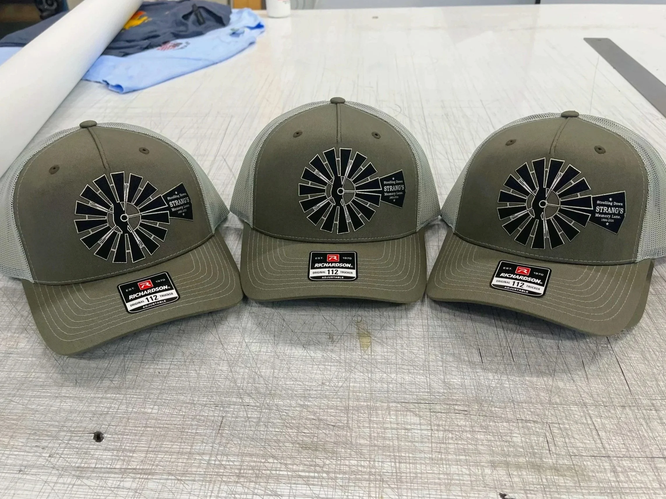 Three green baseball caps with a circular black and silver windmill logo on the front, placed on a worktable in a workshop. The caps have adjustable straps and tags indicating they are Richardson 112 trucker caps.