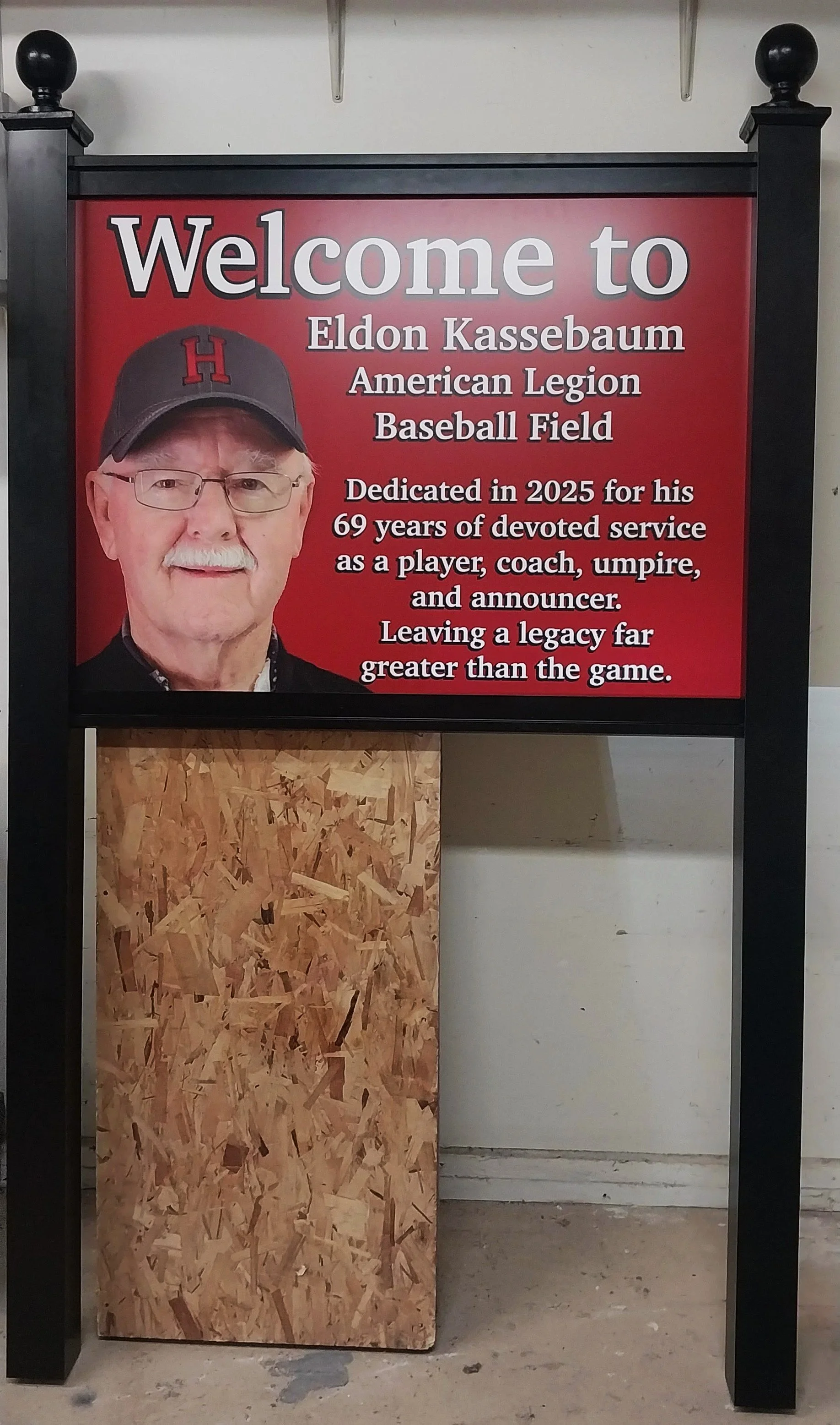 A welcome sign with a photo of Eldon Kassebaum, who is wearing glasses and a baseball cap with a red letter 'H'. The sign is for Eldon Kassebaum at the American Legion Baseball Field, dedicated in 2025 for his 69 years of service as a player, coach, 