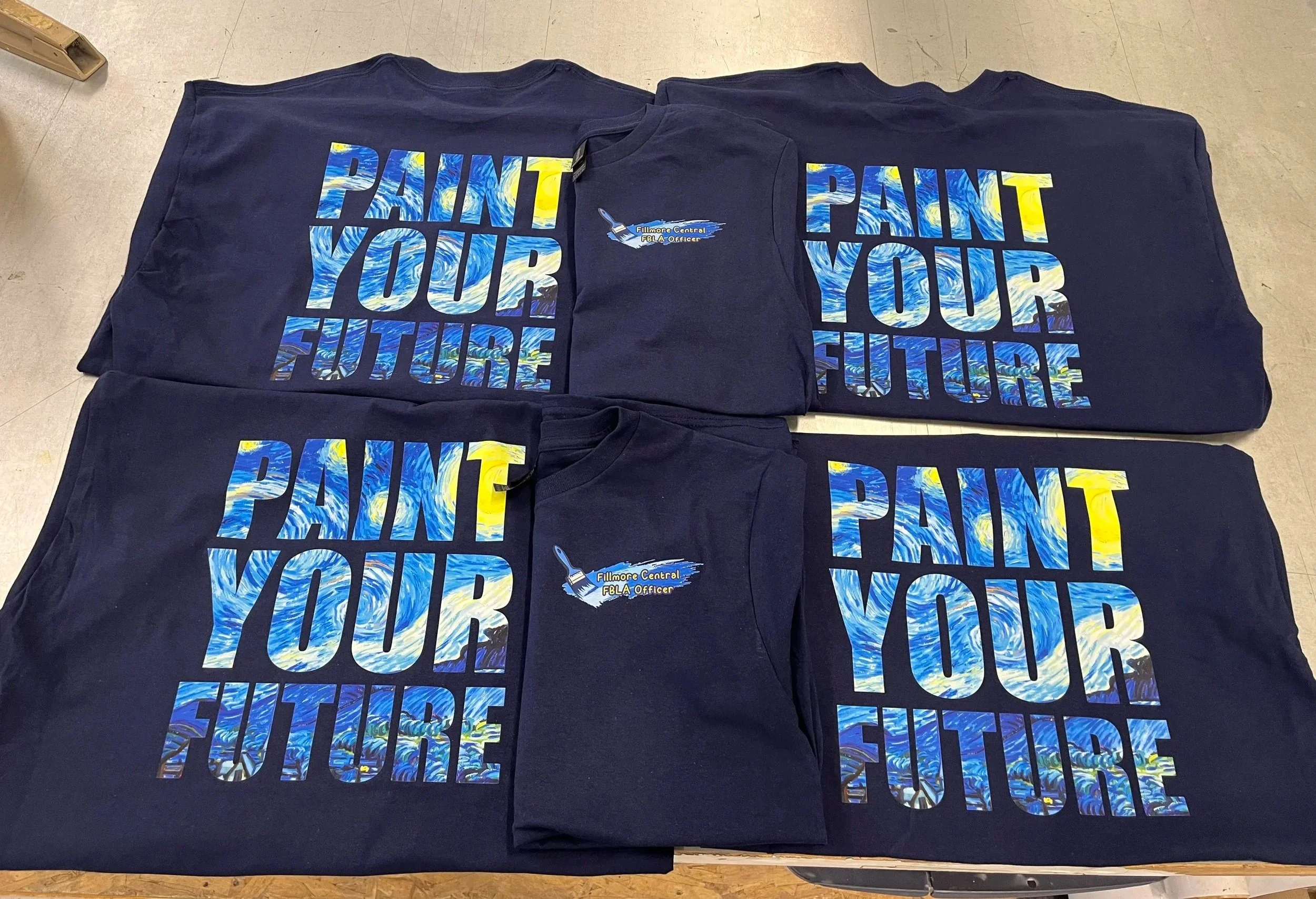 A collection of dark blue T-shirts with a colorful, wave-like design and the phrase 'Paint Your Future' written across the front. Produced by a DTF 