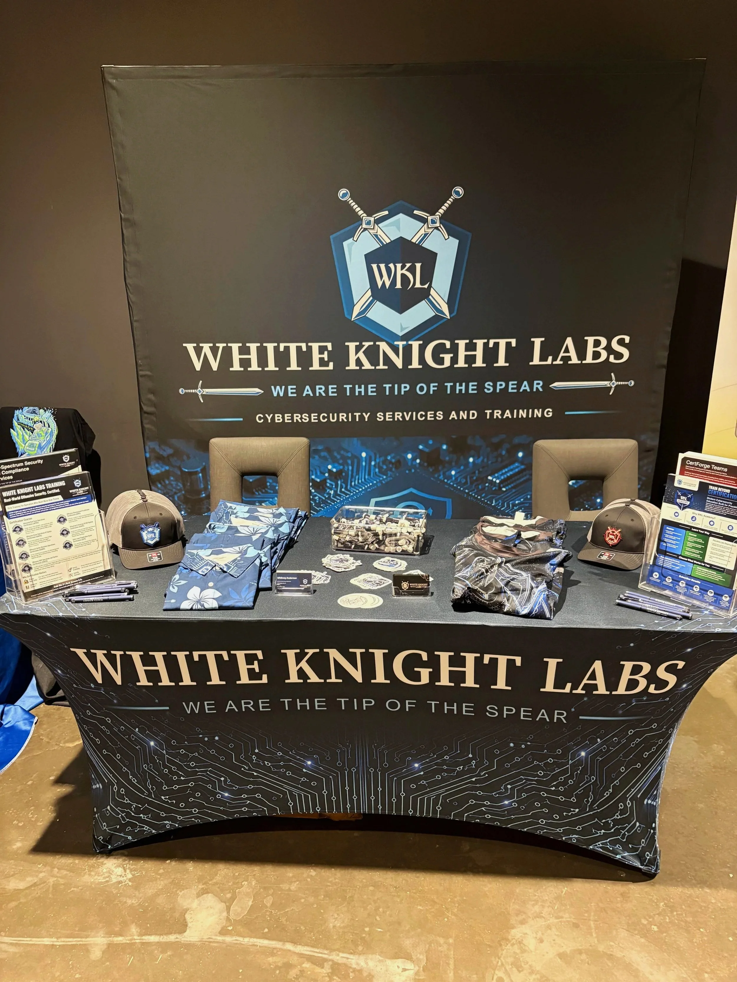 Table display for White Knight Labs with promotional items, informational brochures, hats, and fabric masks, all featuring a tech and cybersecurity theme.
