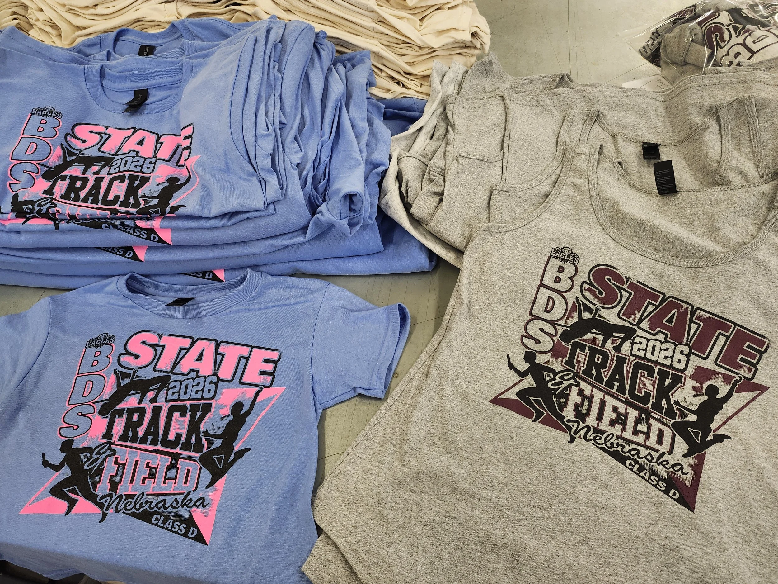 A display of folded T-shirts with school spirit designs for Nebraska Class D sports team, in blue and gray colors, laid out on a surface in a retail or print shop setting.