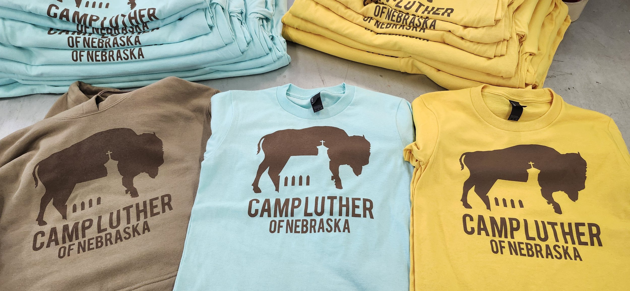 Stacked t-shirts and sweatshirts with a buffalo graphic and the text 'Camp Luther of Nebraska' in blue, brown, and yellow colors.