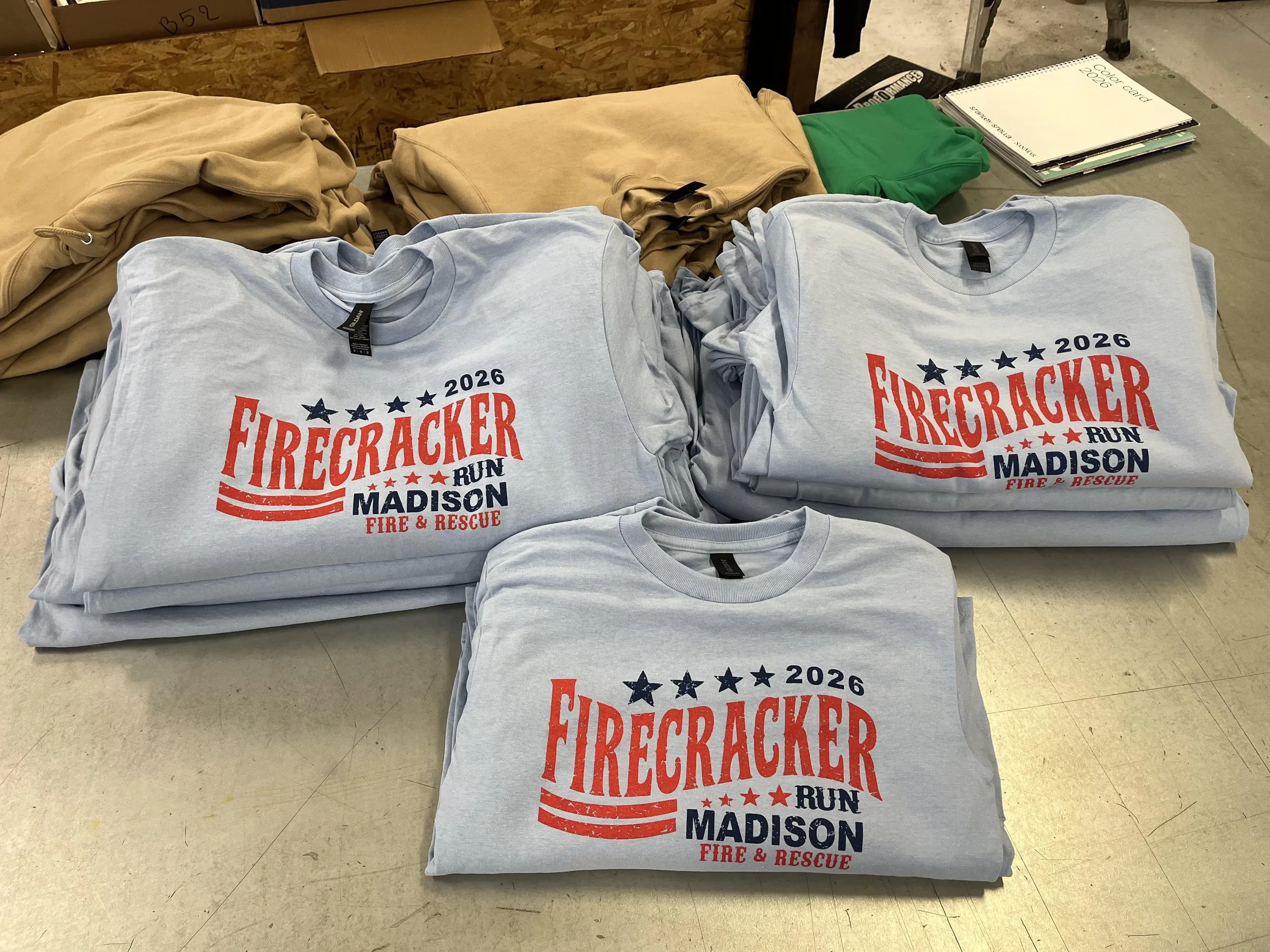 Stacks of beige and white T-shirts and a green hoodie on display table, some with a red, black, and blue Firecracker Run Madison logo and text, in an indoor setting.