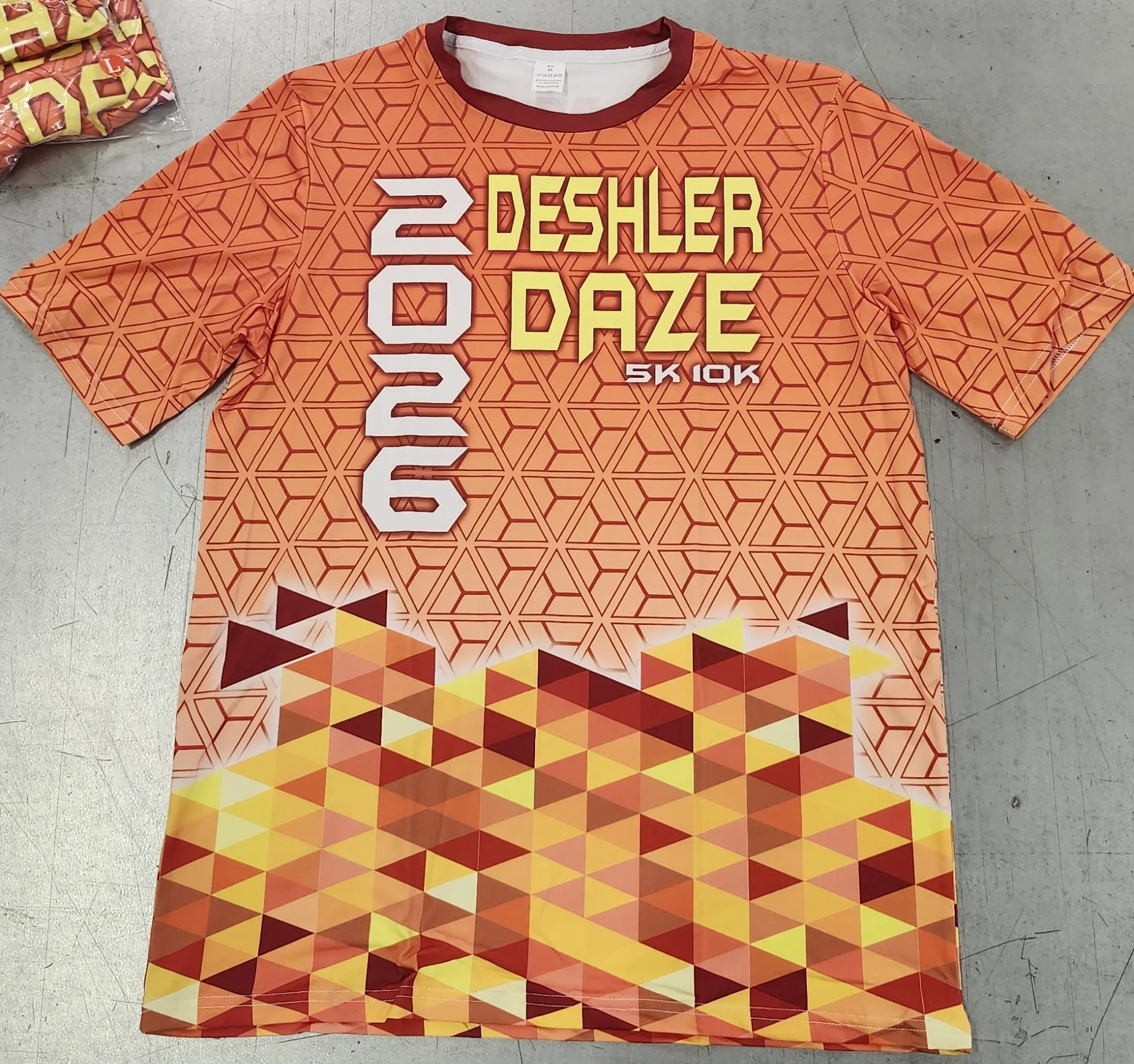 Orange Sublimated T-shirt with geometric pattern and bold text that reads '2026 Deshler Daze 5K 10K' layered over the pattern.