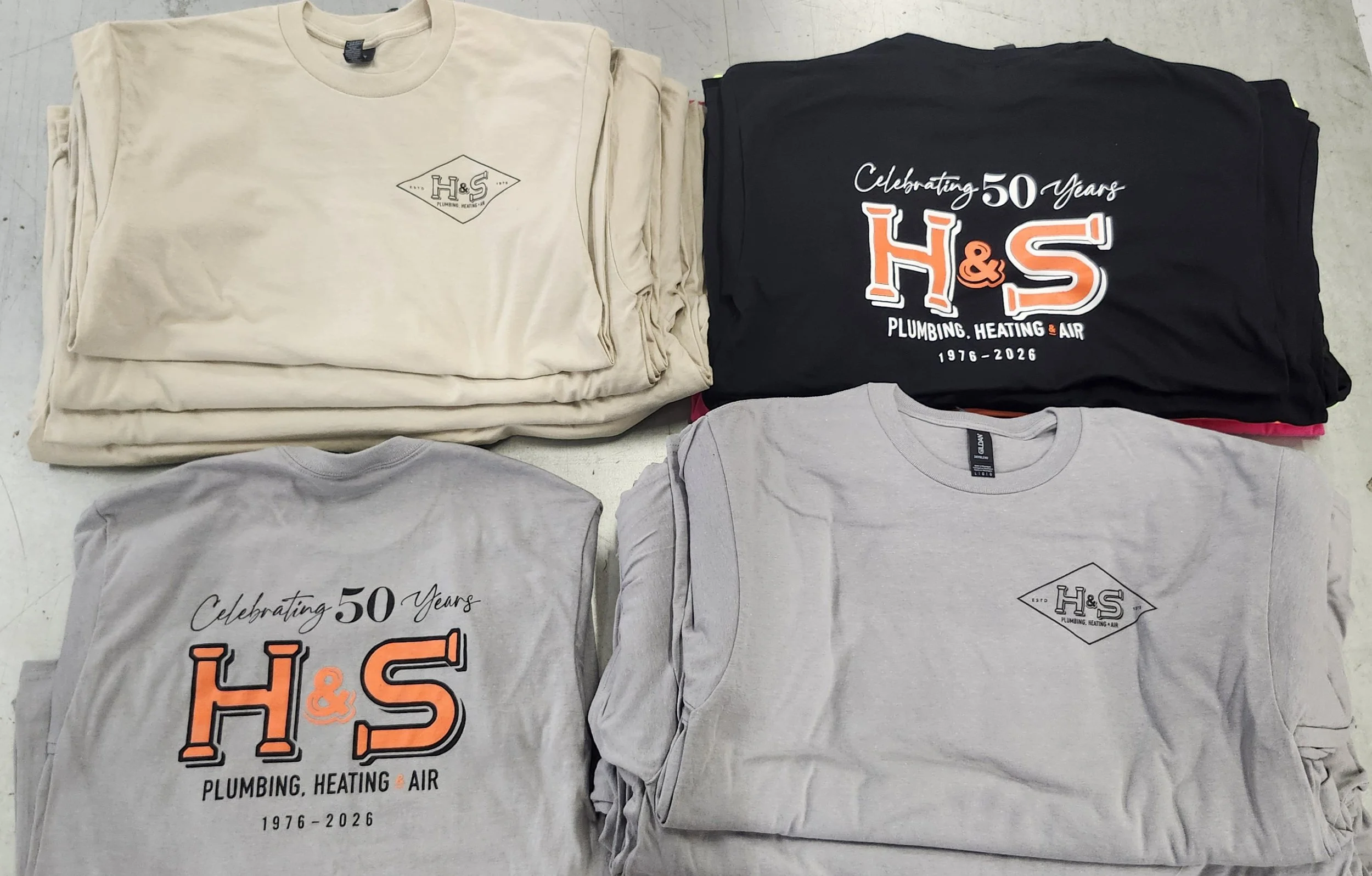 Several stacks of screen-printed t-shirts, with two notable designs in dark and light colors. The black shirt features a logo celebrating 50 years of a plumbing, heating, and air company from 1976 to 2026. The light-colored shirts display text celebr