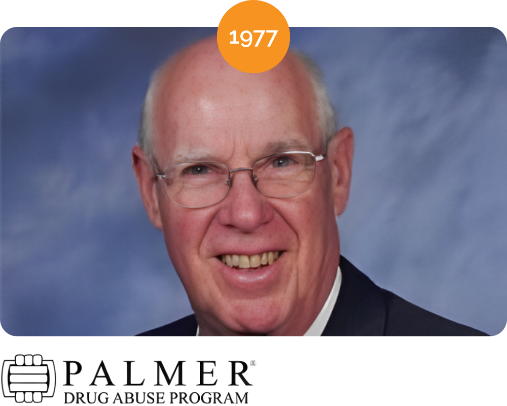 Portrait of a smiling middle-aged man with glasses, wearing a suit, set against a blue background, with a 1977 banner at the top.