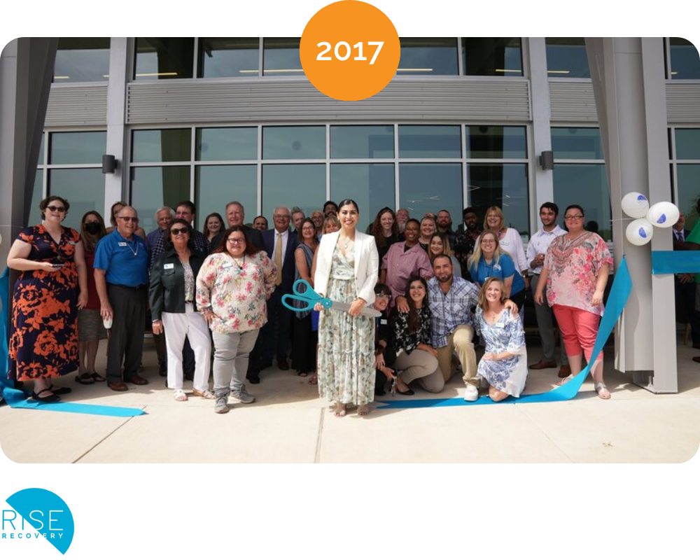 Group of people celebrating a ribbon-cutting event in front of a modern building, with a woman holding large scissors. The event took place in 2017, as indicated by the orange circle at the top. There is a blue ribbon on the ground and white balloons on the right side.