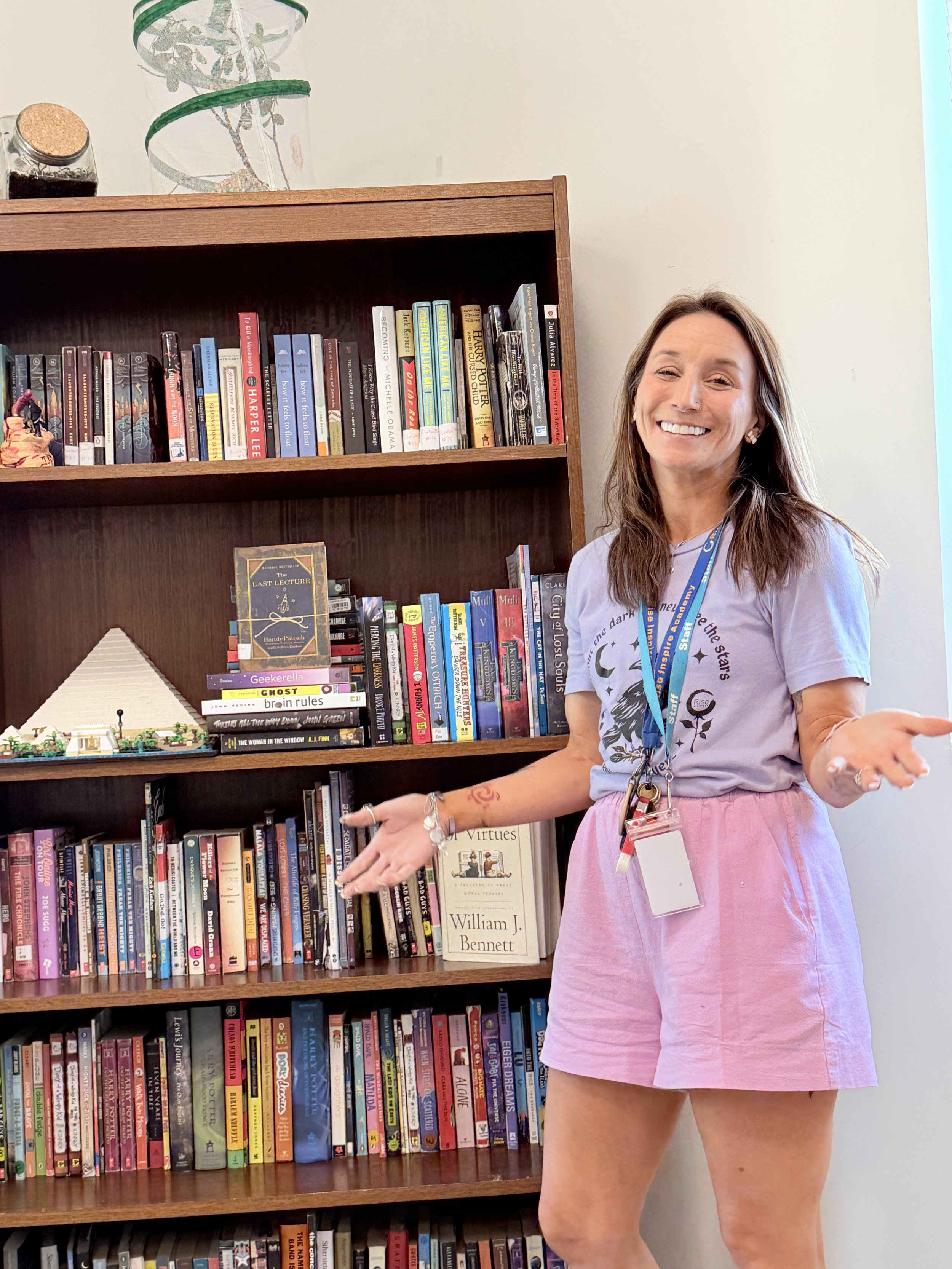 A woman smiling and gesturing with her hands in front of a bookshelf filled with various books, a Lego display, and a poster of William J. Bennett.
