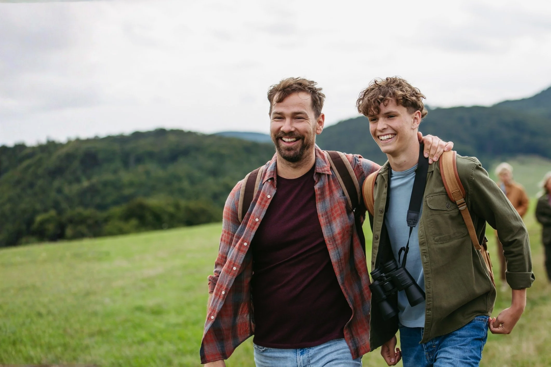A man and a boy smiling, walking outside in a green field with hills in the background, both wearing backpacks.