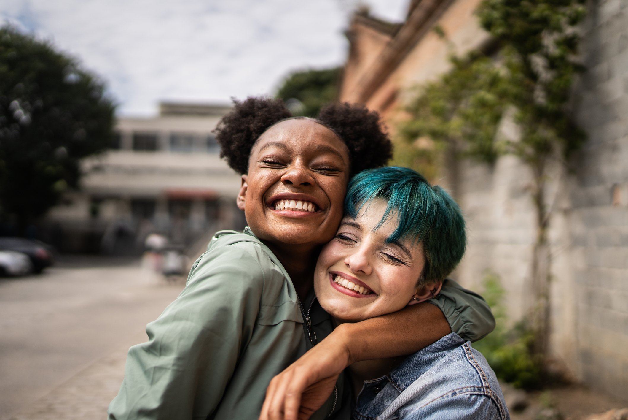 Two women hugging and smiling outdoors with trees and buildings in the background.