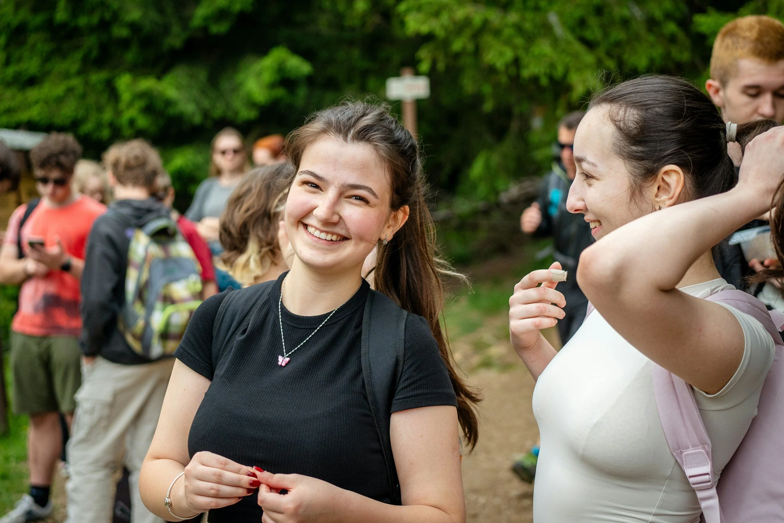 Smiling young woman with long dark hair wearing a black t-shirt and a butterfly necklace standing outdoors surrounded by a group of people, some of whom are looking at their phones and some are talking, with lush green trees in the background.