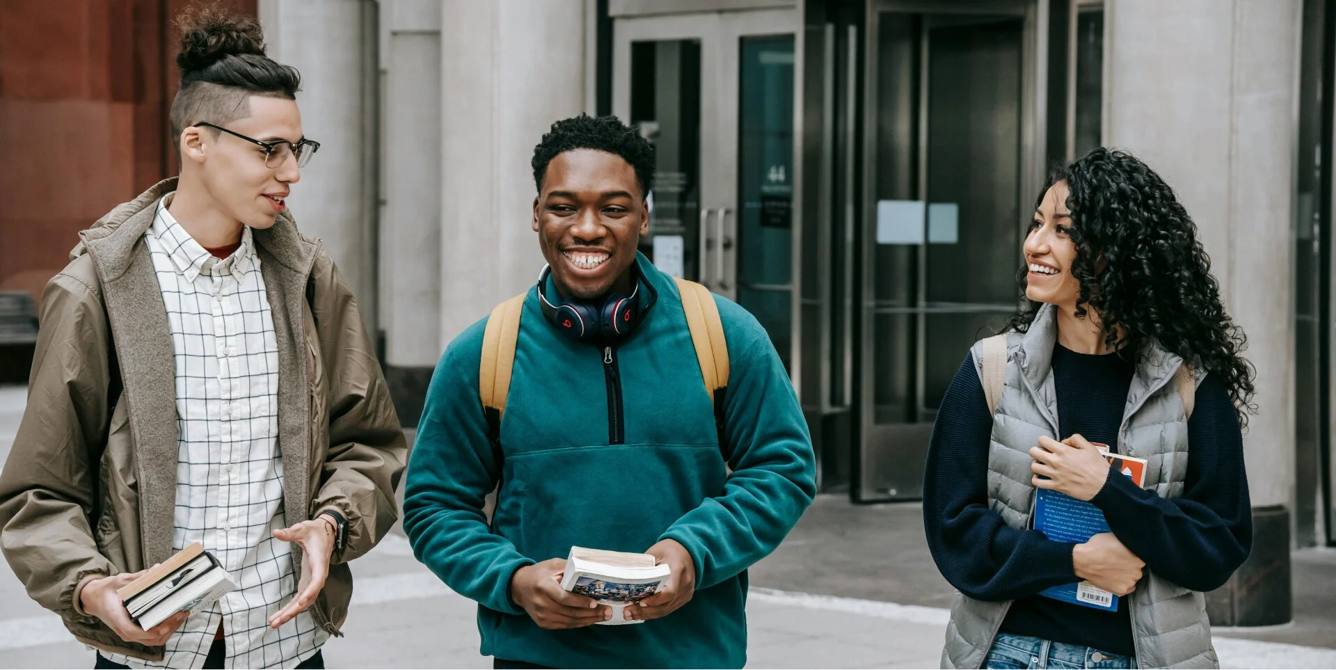 Three young adults standing on a city sidewalk, engaged in conversation and smiling. The person in the middle is wearing a teal jacket and headphones around his neck, holding books. The person on the left is wearing glasses and holding a small stack of books. The person on the right has curly black hair and is holding a notebook or folder, wearing a gray vest over a black sweater.