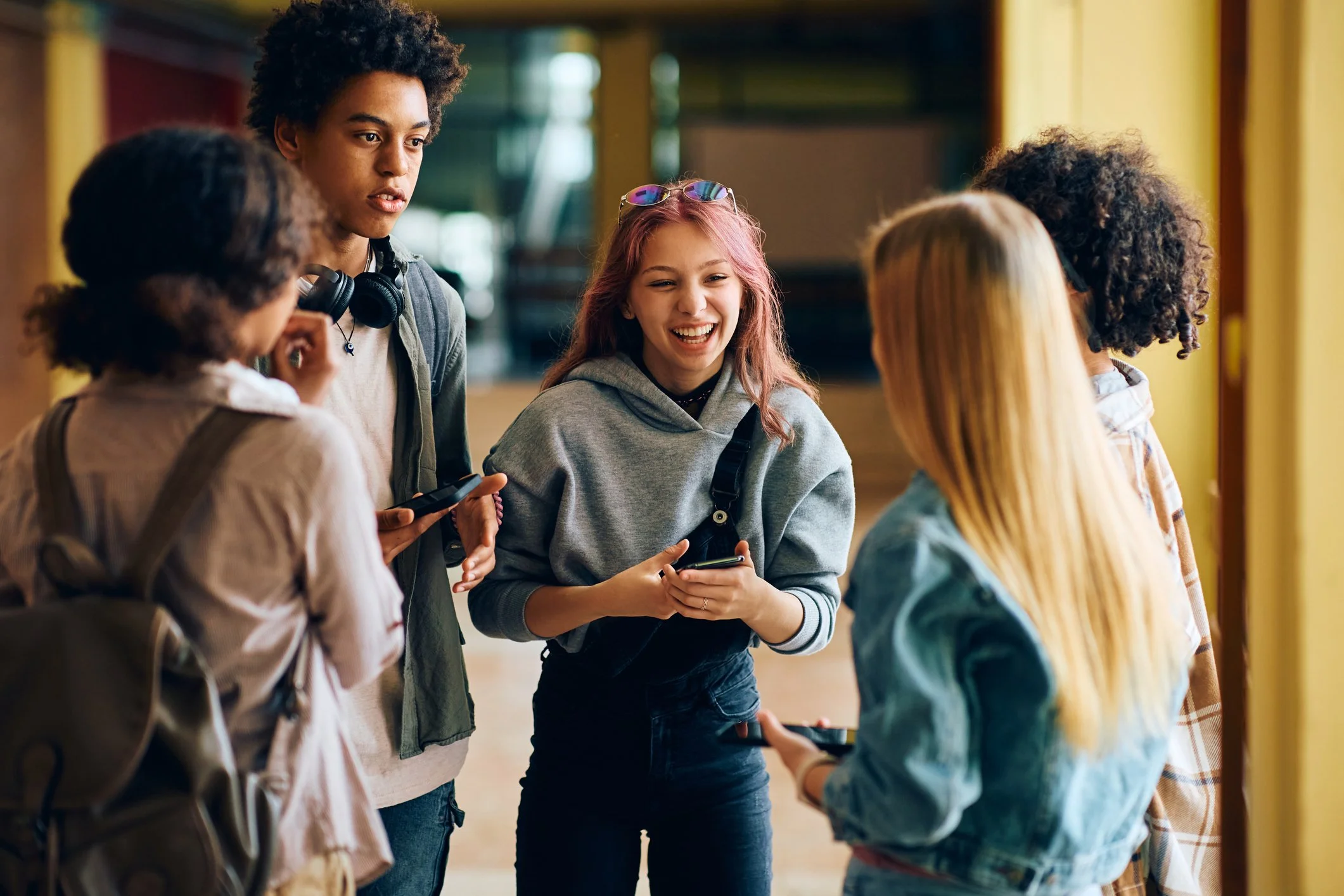 A group of five teenagers having a conversation indoors, some holding smartphones, with a yellow structure on the right and a background of blurred green and yellow elements.