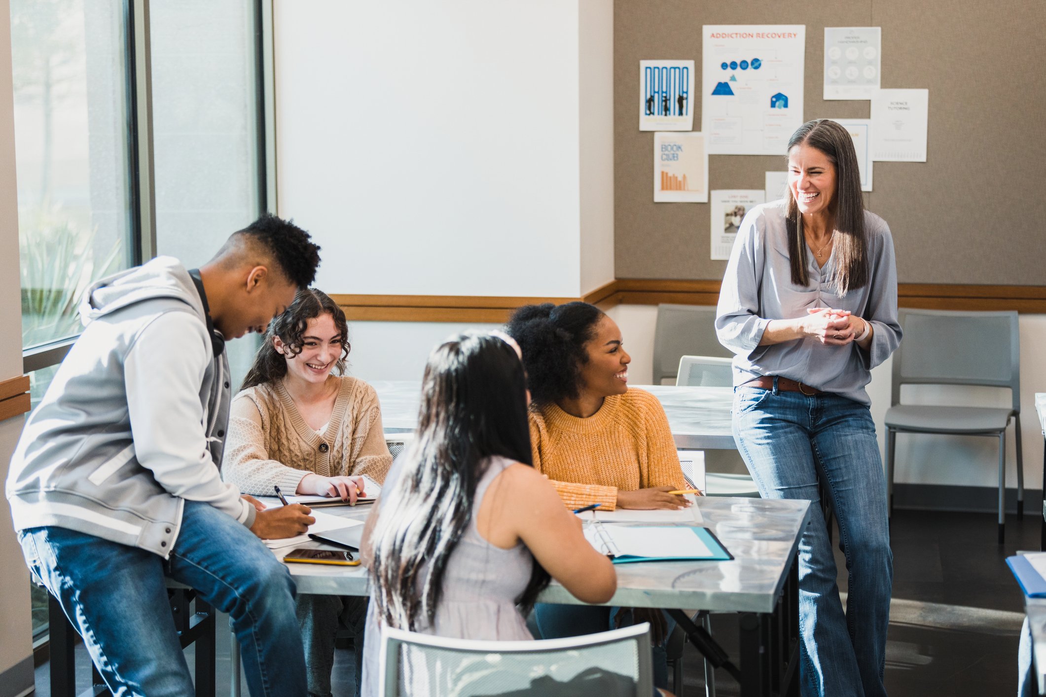 A diverse group of students sitting at a table in a classroom, listening to a smiling woman standing near the wall, who appears to be teaching or leading a discussion. The classroom has posters on the wall, including one about addiction recovery, and large windows letting in natural light.