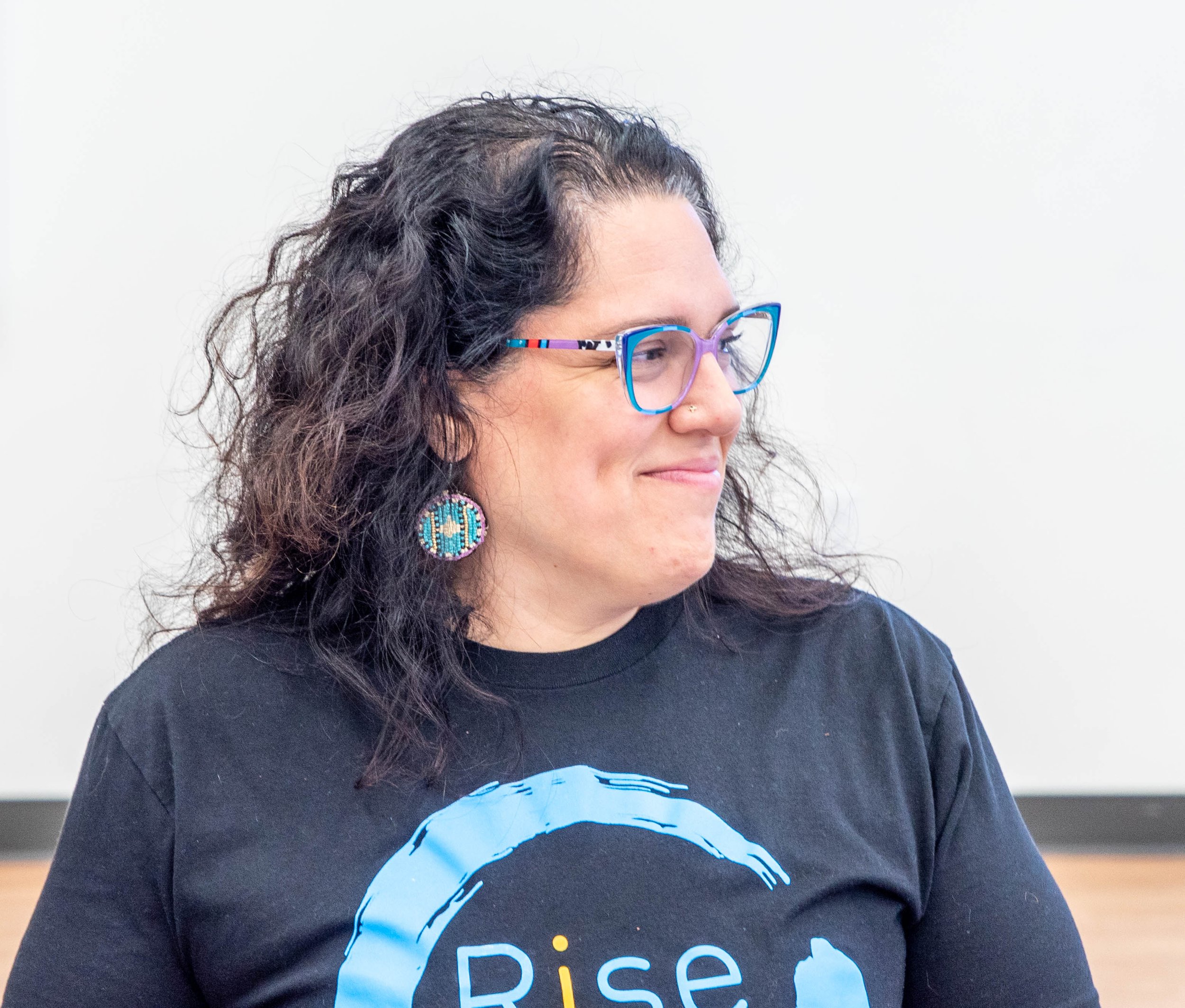 Profile of a woman with curly dark hair, wearing colorful glasses, earrings, and a black T-shirt with a blue and white circular design