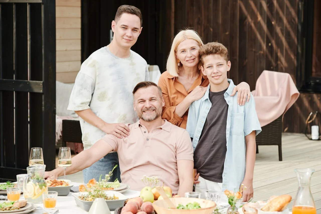 A family of five gathered on a deck for a meal, smiling and posing for the photo, with food and drinks on the table.