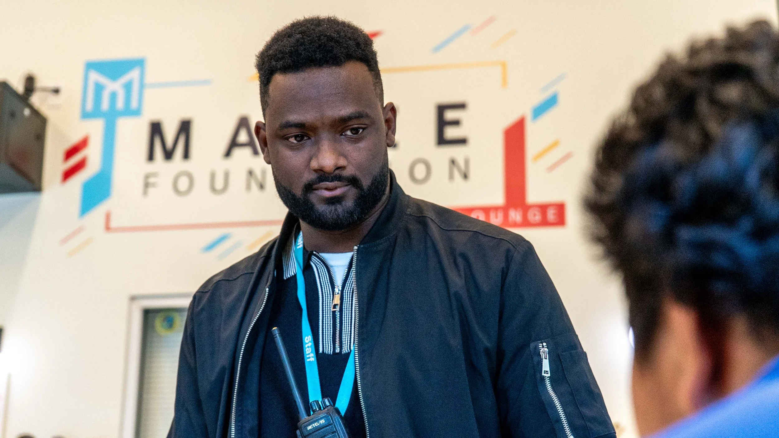A man with a beard and short hair standing indoors, wearing a black jacket and a lanyard with an ID badge, is looking at a person in front of him. There is a colorful banner in the background with the words 'MARE FOUN' visible.