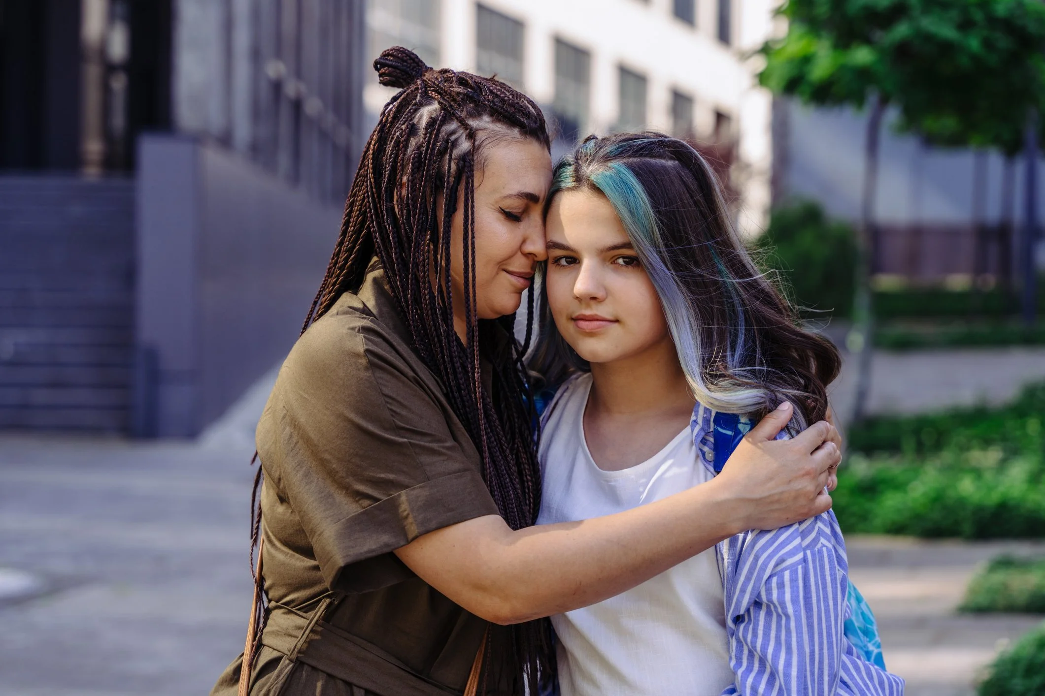 Two women sharing an embrace outdoors, one with braided hair and the other with multicolored hair, standing on a city sidewalk with buildings and trees in the background.