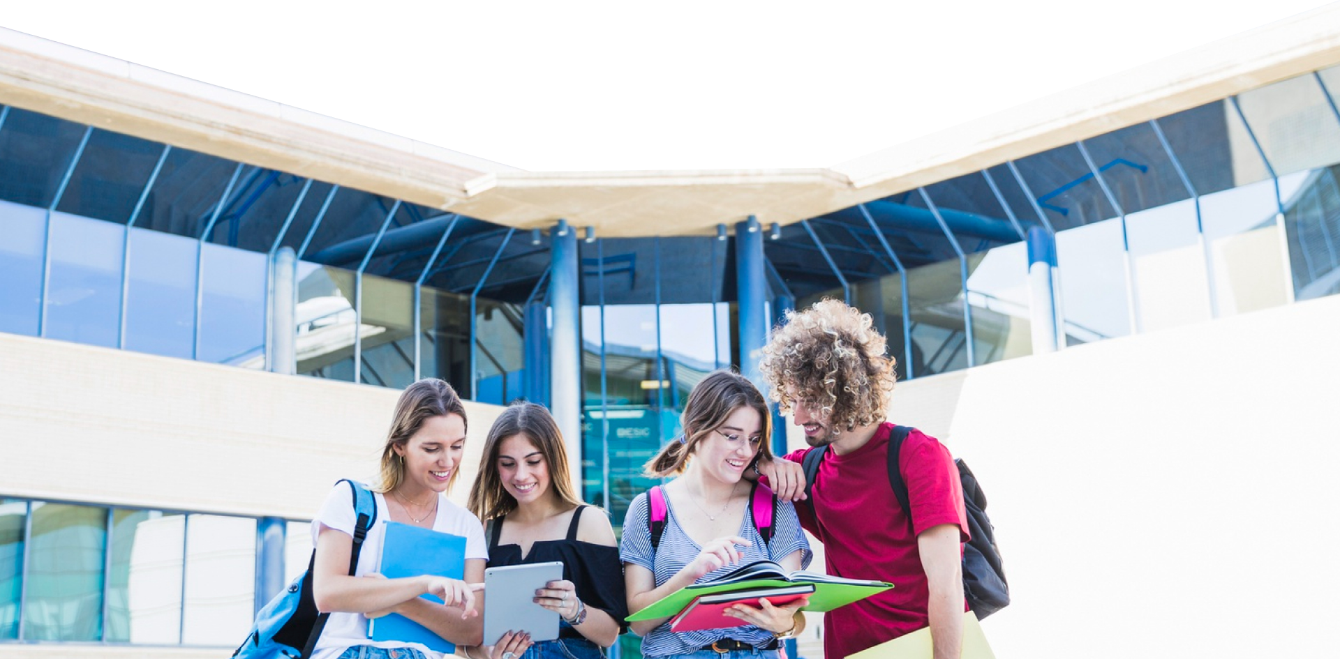 Group of four college students studying outside a modern glass building, all smiling and looking at books and tablets.