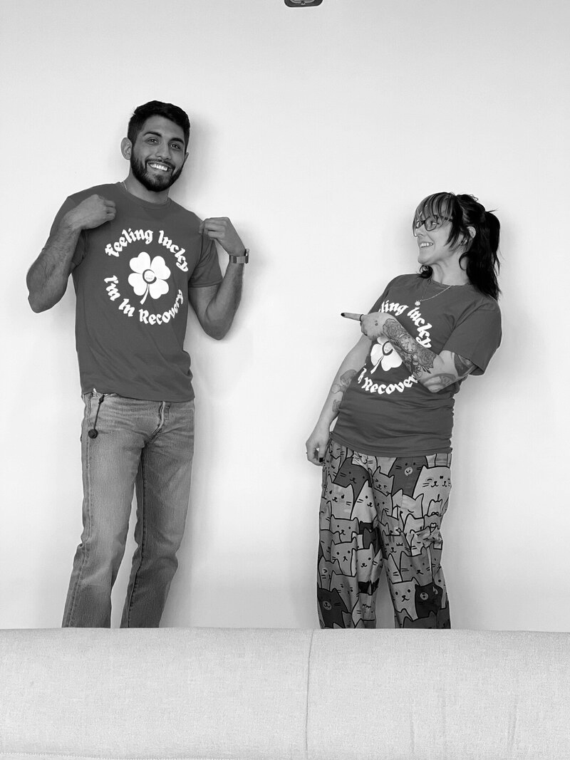 Two people, a man and a woman, standing against a plain wall, both wearing matching T-shirts with a four-leaf clover and the text 'Feeling Lucky, I'm in Recovery.' The man is smiling, with his hand on his chest, while the woman is pointing at him and smiling.