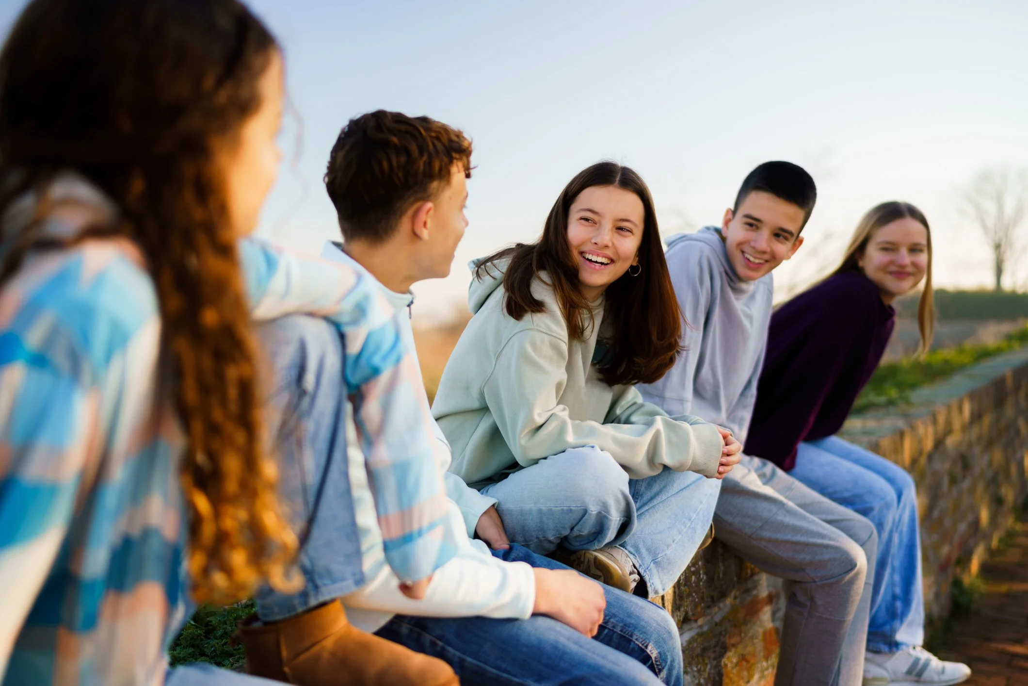 Group of five teenagers sitting on a low wall outdoors, chatting and smiling during sunset.