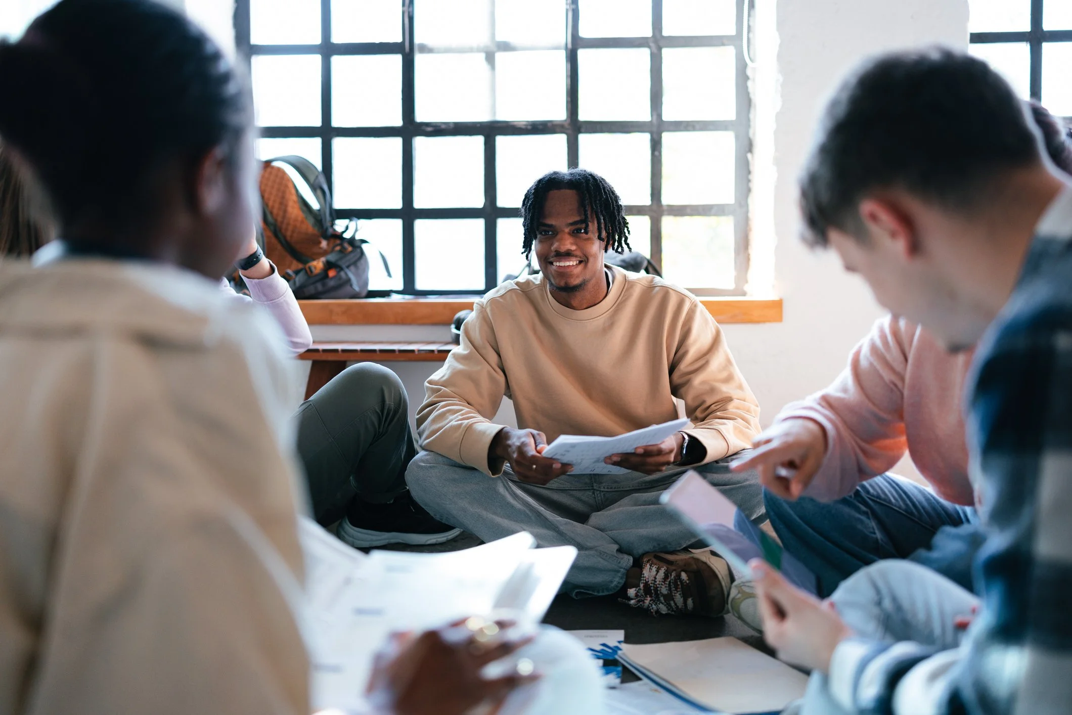A group of young people sitting on the floor in a circle, engaged in conversation with a smiling man holding papers, near a large window with grid panes.