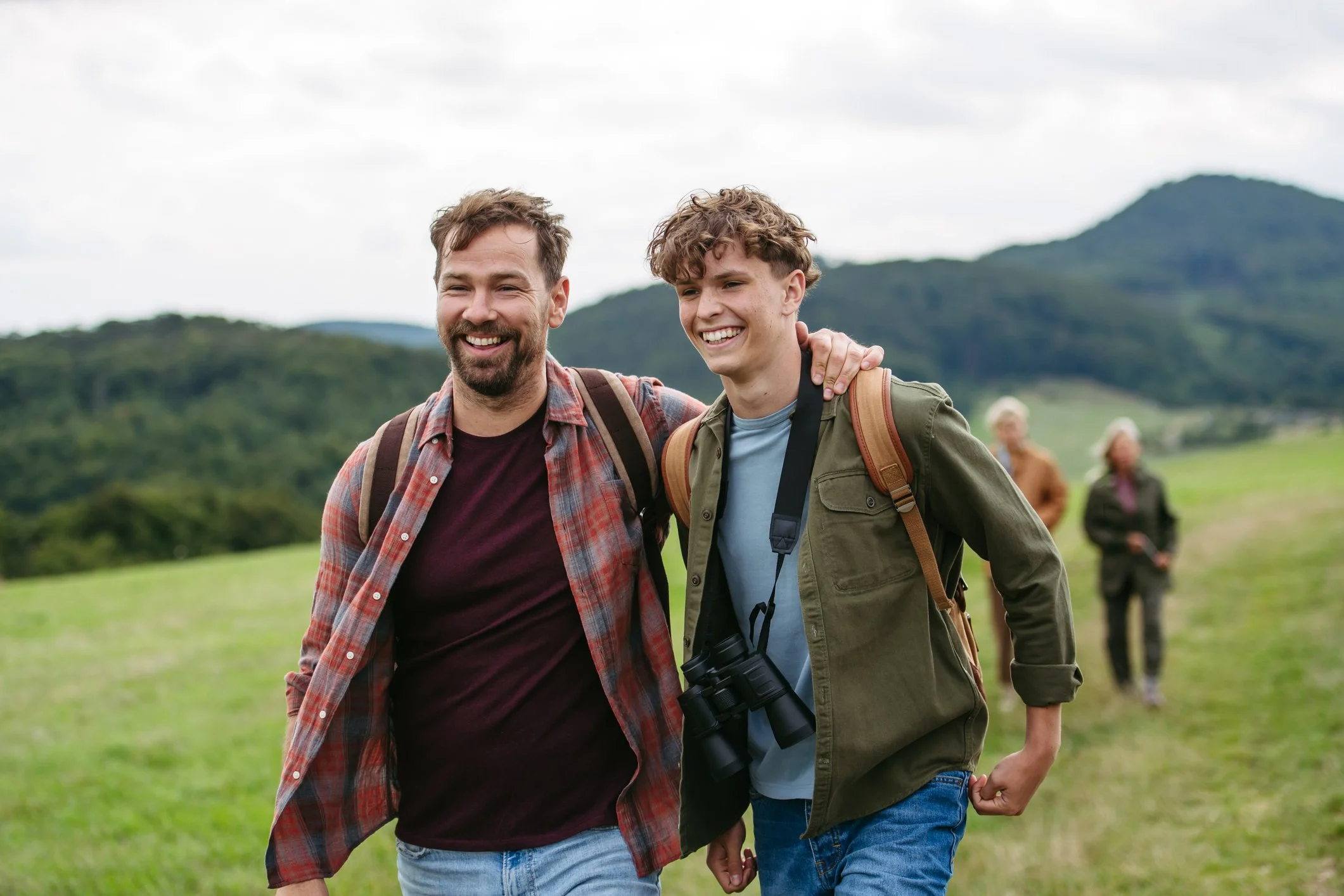 Two men walking outdoors in a grassy area with hills in the background, smiling and holding hands.