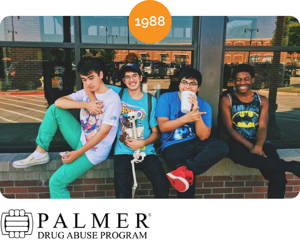 Four teenage boys sitting on a brick ledge outside a building with large windows, smiling and interacting, dressed in colorful casual clothes from 1988.