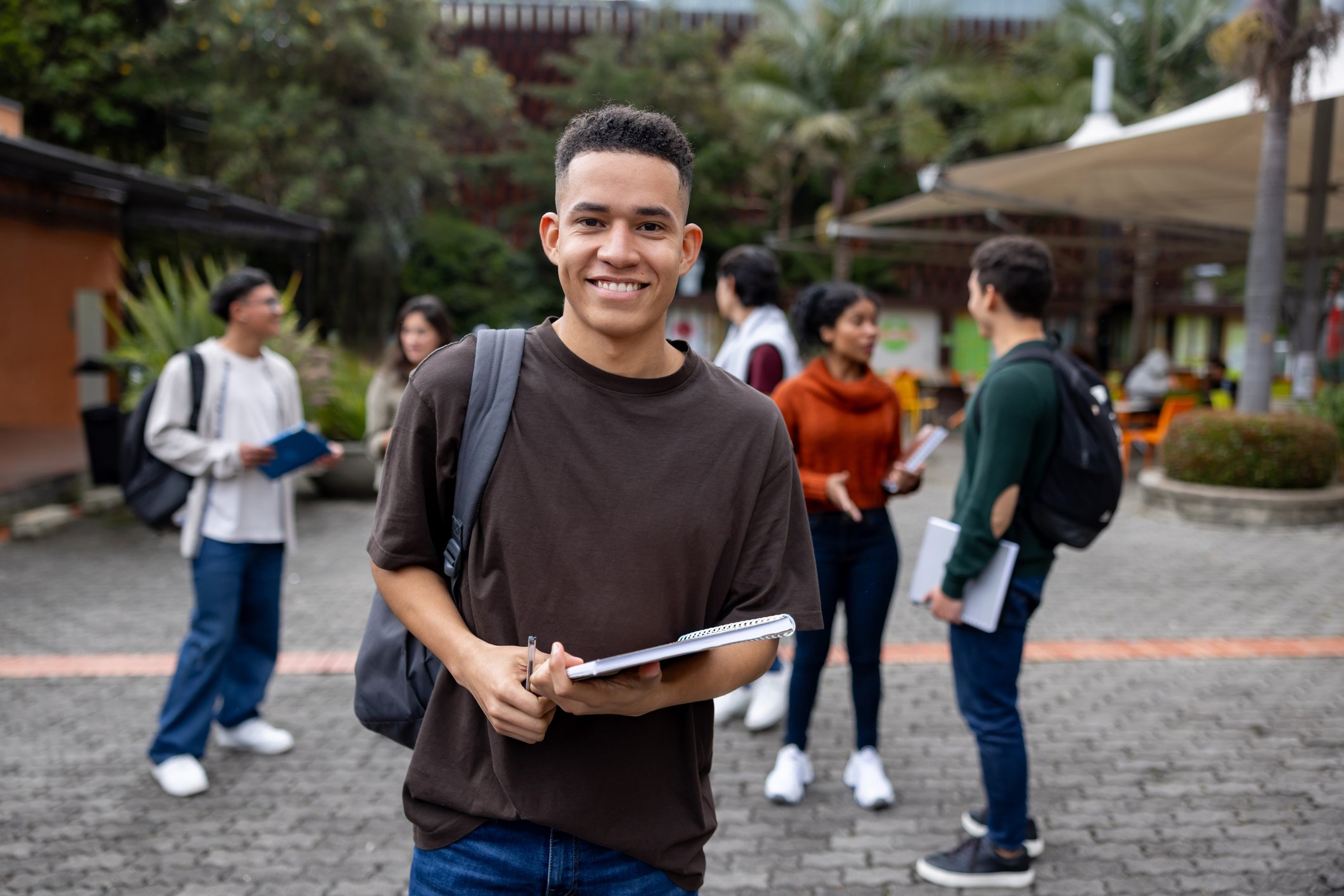 Young man with short curly hair, smiling, standing outdoors with a group of people in the background. He is holding a notebook and a pen, wearing a brown T-shirt and a backpack. The background shows trees and a courtyard area.