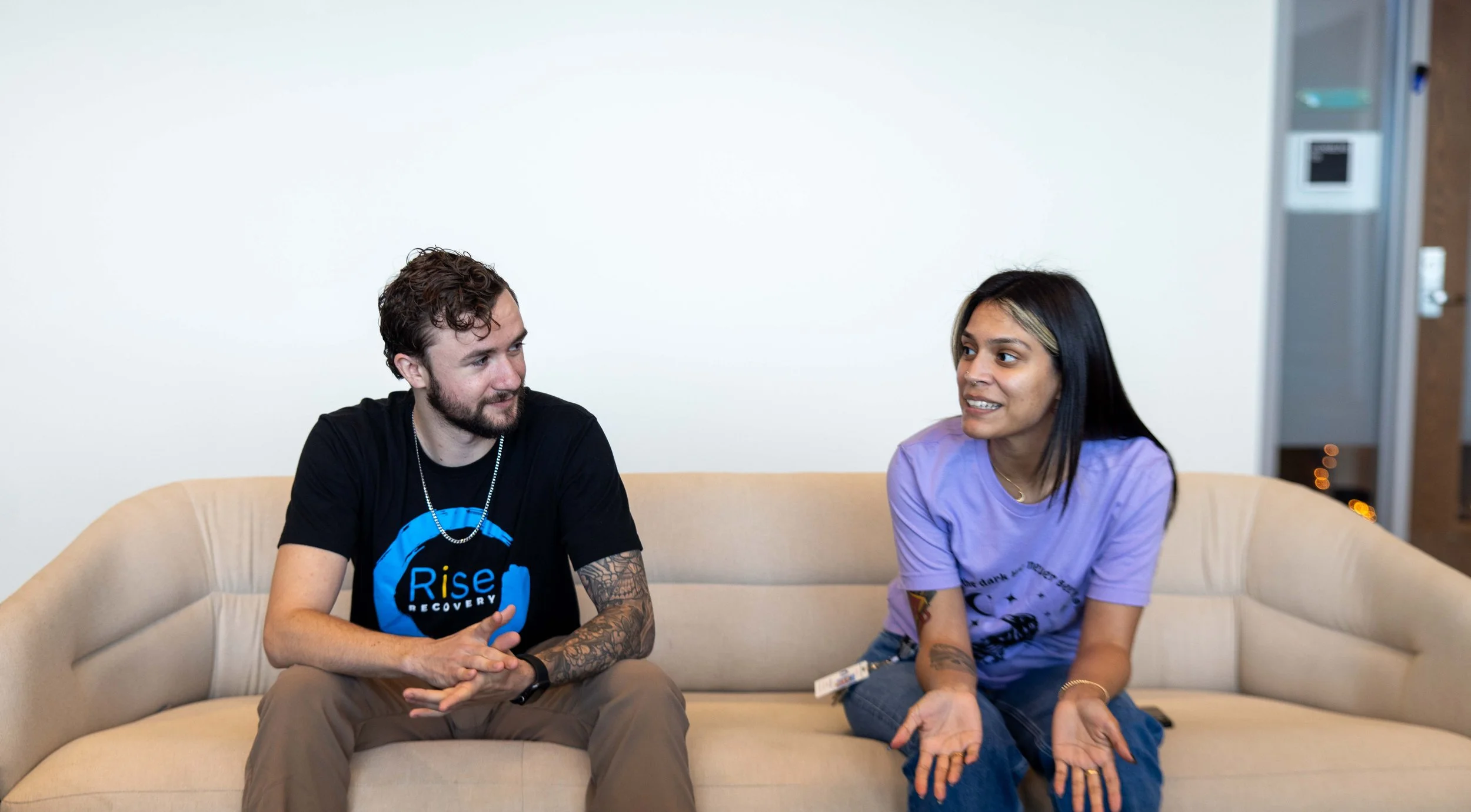 Two people sitting on a beige couch engaged in conversation. The man on the left has curly hair, a beard, and tattoos on his arms, wearing a black T-shirt with a blue logo. The woman on the right has long black hair with blonde highlights, wearing a purple T-shirt, and gesturing with her hands.