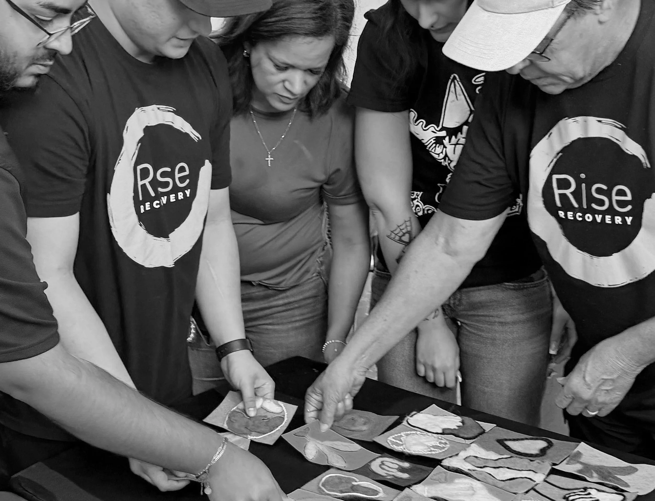 Group of people gathered closely together, engaging in an activity involving colorful cutouts or cards on a table, with some wearing t-shirts with the text 'Rise Recovery'.