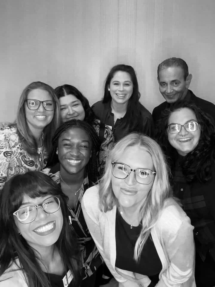 A group of nine diverse people, mostly women, smiling and posing together indoors.