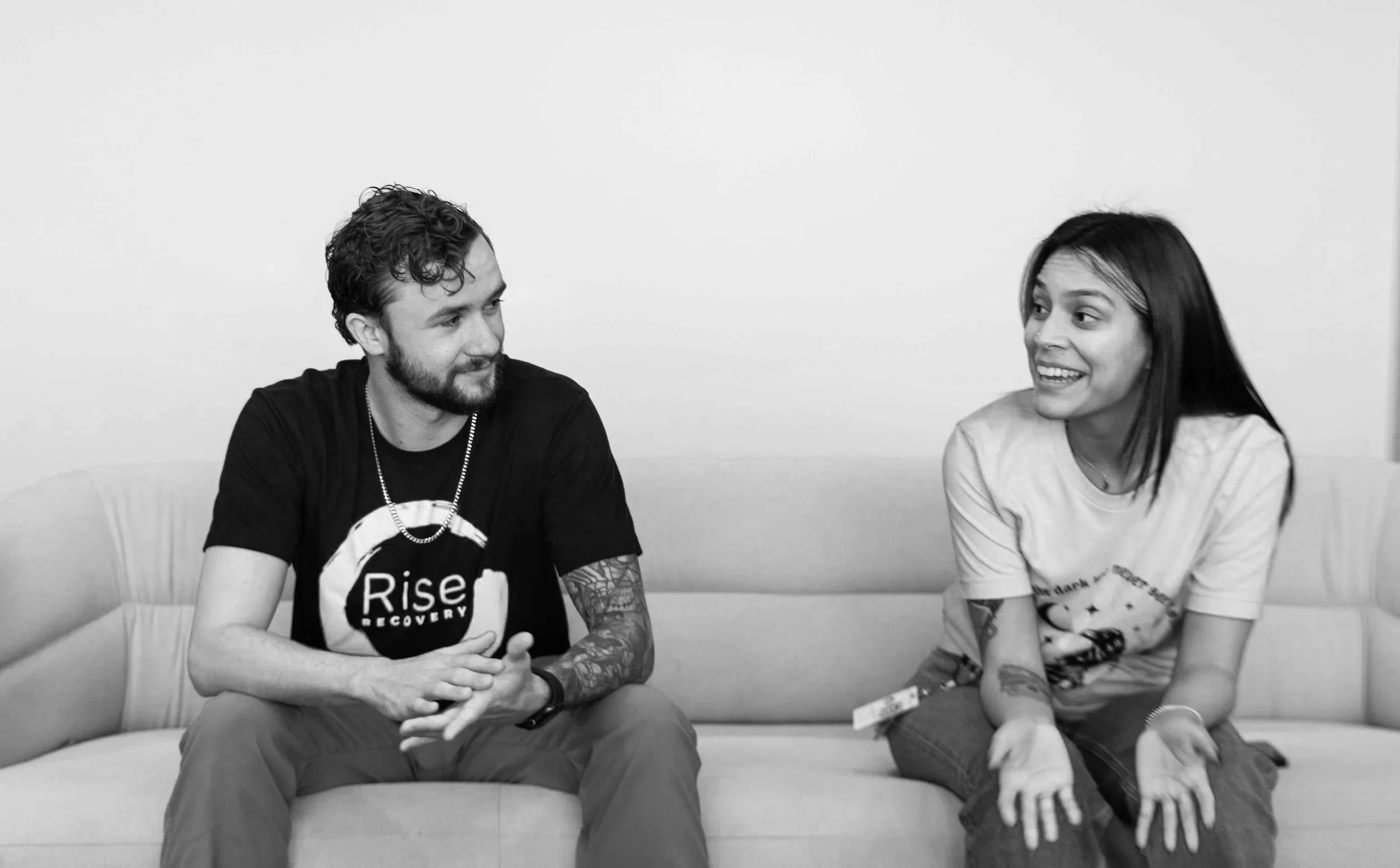 A black and white photo of a man and a woman sitting on a sofa and talking to each other. The man has a beard, curly hair, tattoos on his arms, and is wearing a T-shirt with a logo. The woman has long dark hair, is smiling, and is wearing a T-shirt with printed text. They appear engaged in conversation.