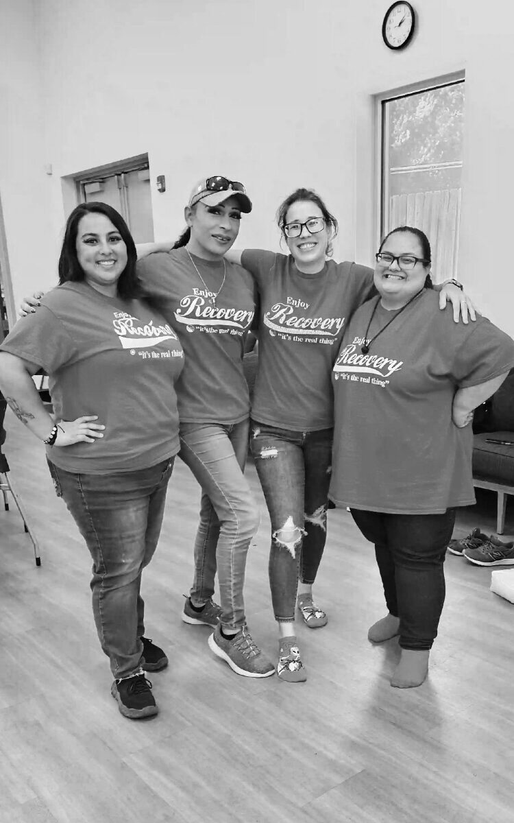 Four women standing together indoors, smiling and posing for a photo, wearing matching t-shirts that say 'Enjoy Recovery - It's the real thing.'