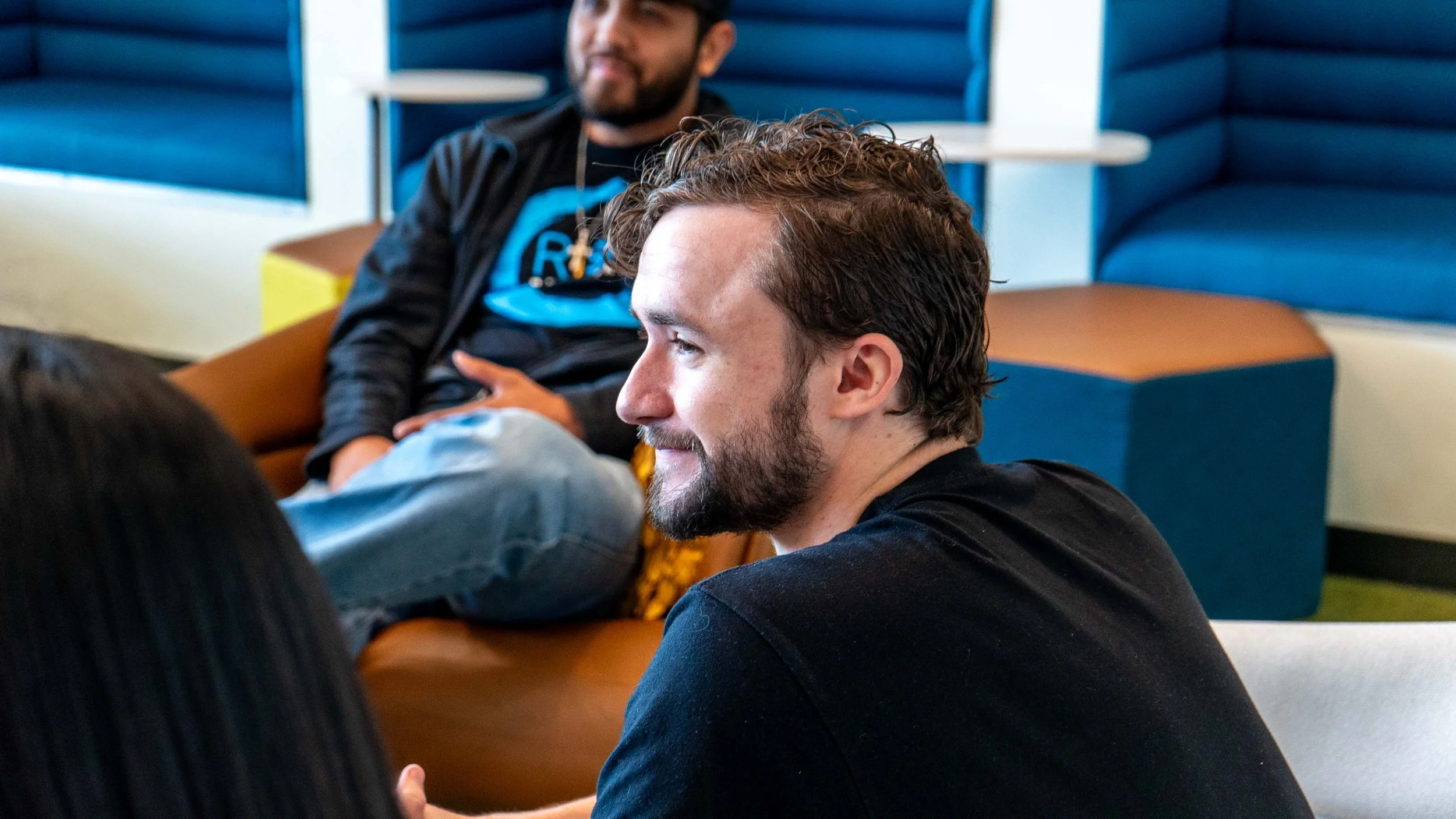 Two men sitting at a table in a casual setting, engaged in conversation, with colorful booth seating in the background.