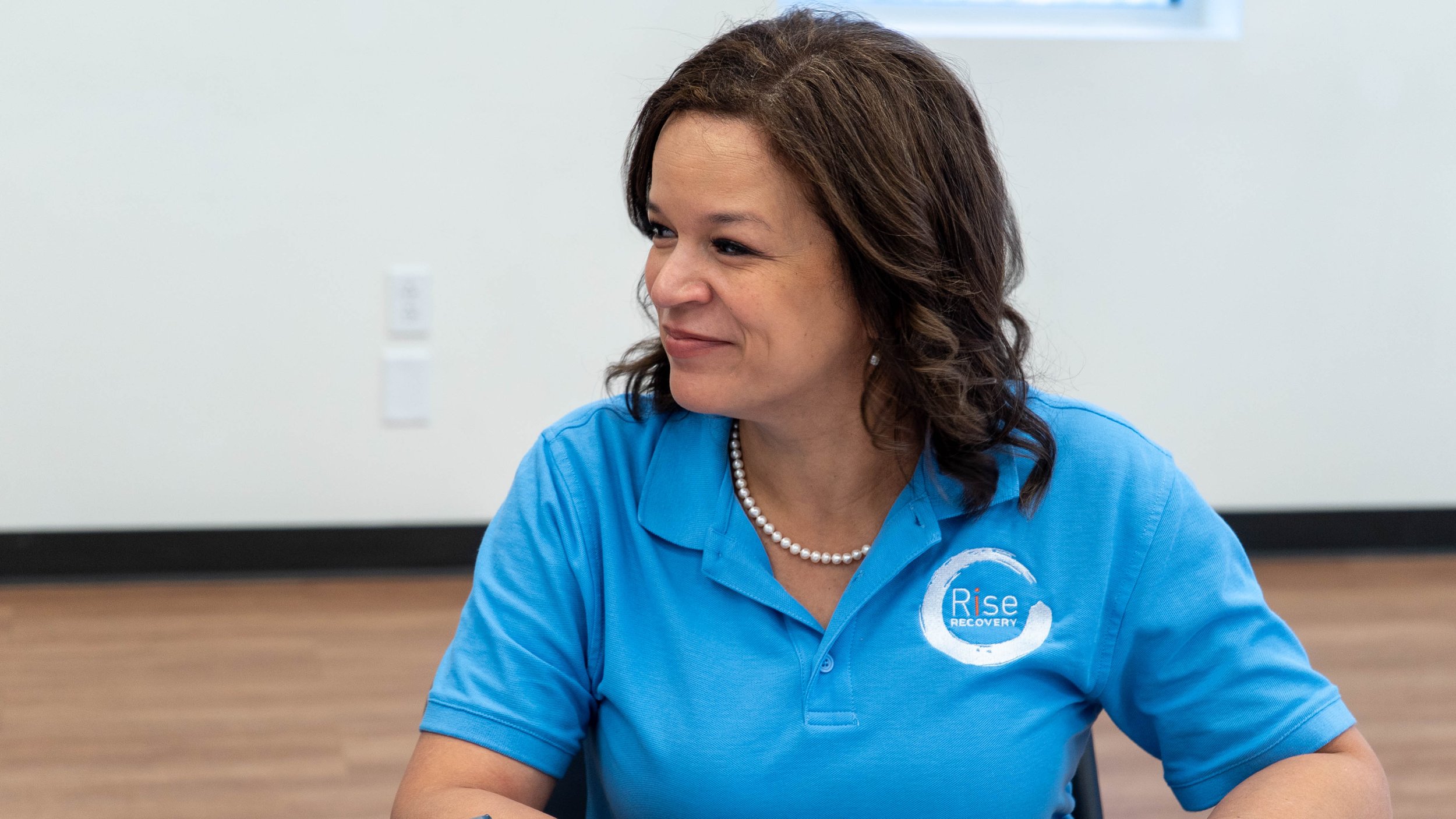 A woman with brown hair wearing a blue polo shirt with 'Rise Recovery' logo, sitting at a table in a room with white walls, smiling.