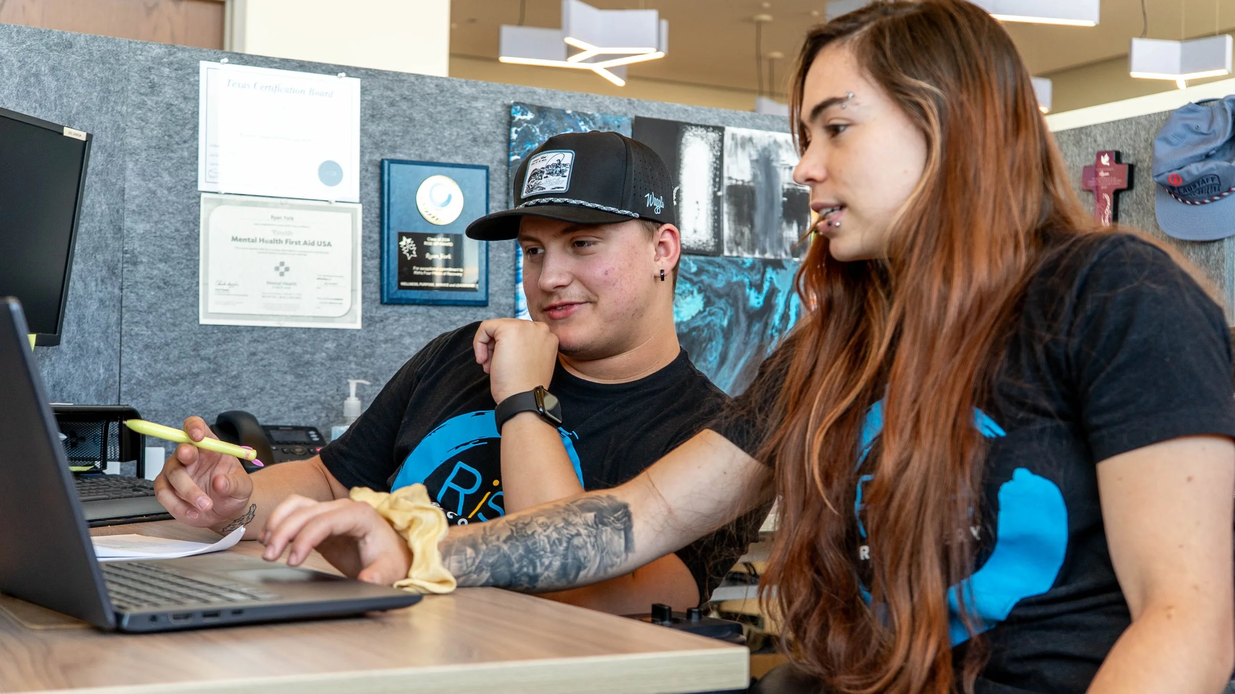 Two people sitting at a desk, looking at a computer screen in an office setting, with framed awards and artwork on the wall behind them.