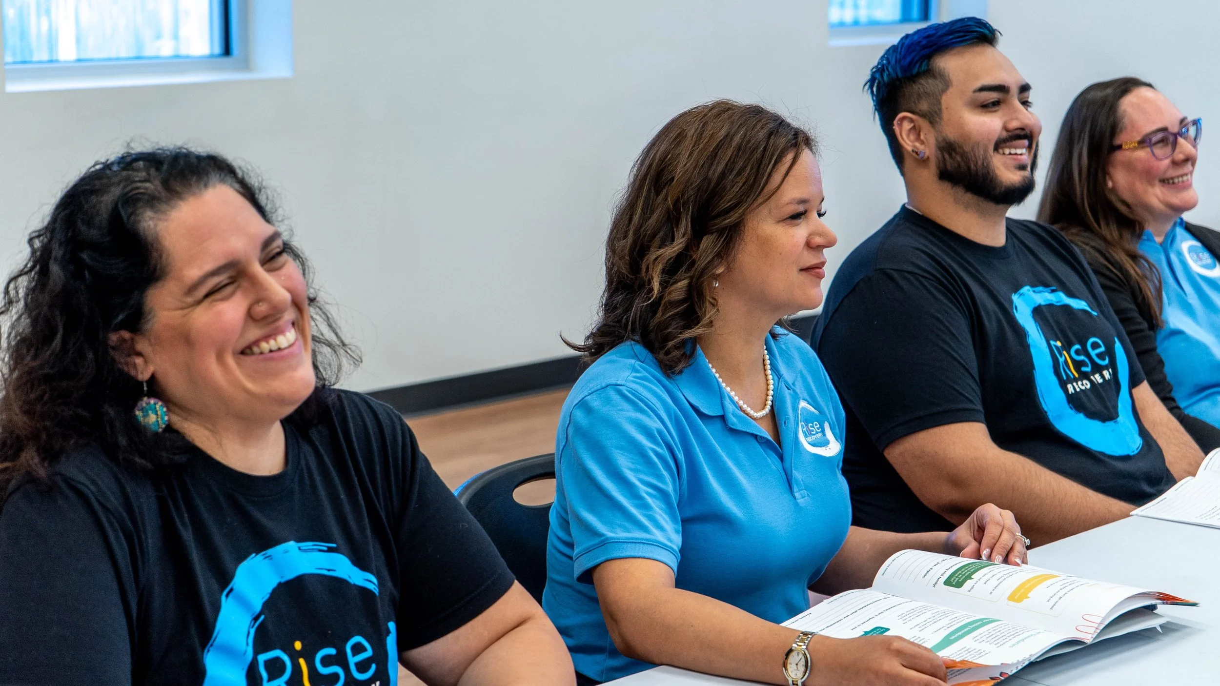 A diverse group of four smiling people attending a meeting, sitting at a table with open books, wearing black or blue Rise T-shirts or polo shirts.